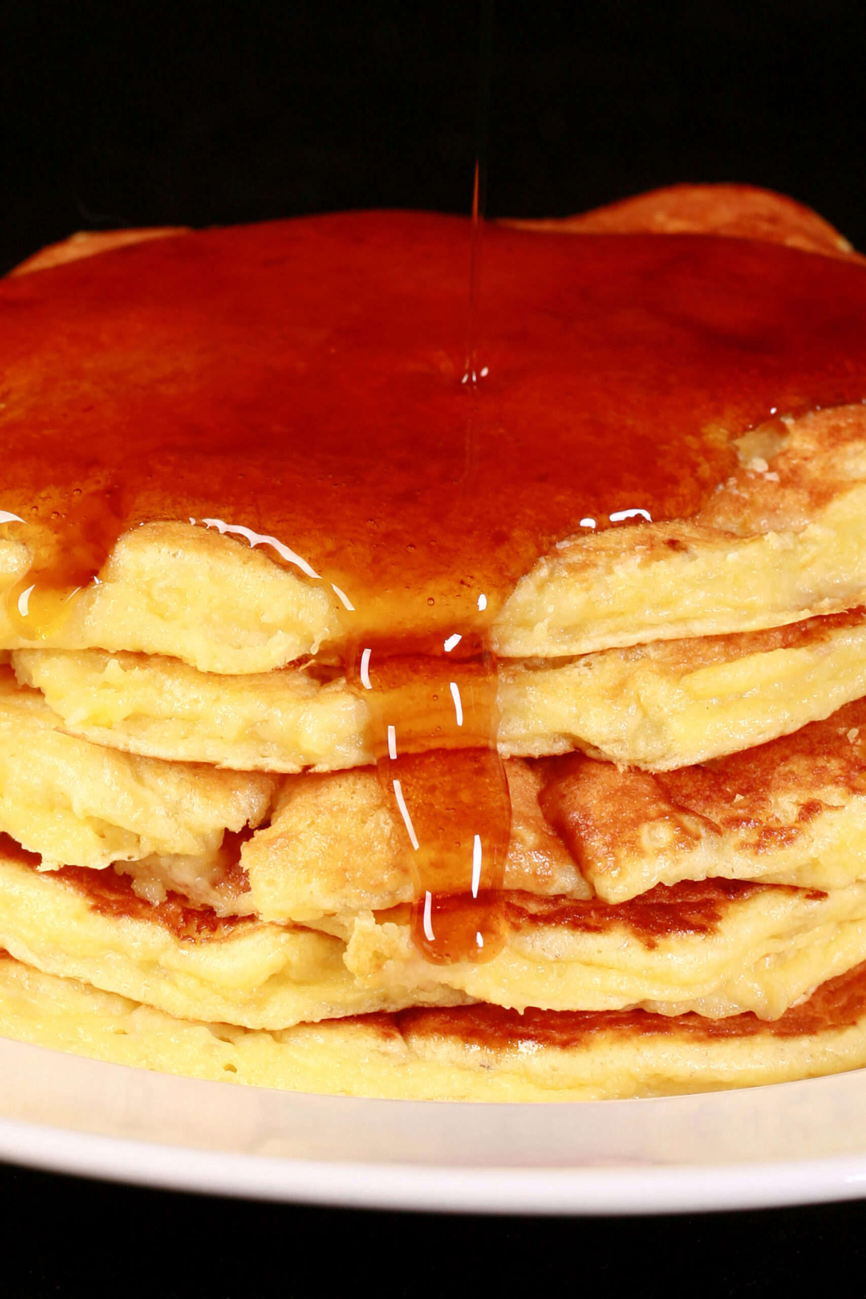 A stack of 6 banana protein pancakes on a place, with maple syrup tripping off the top.