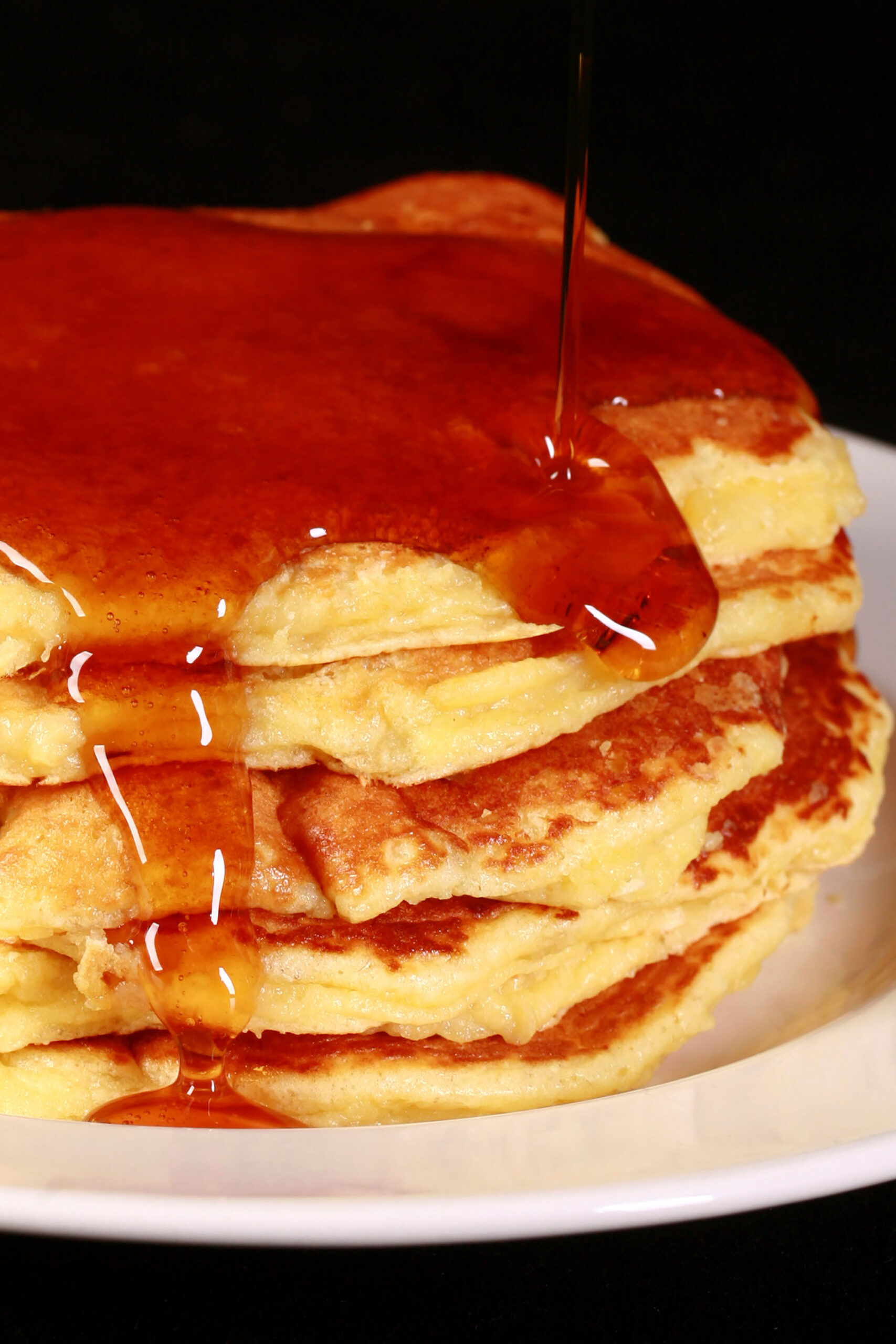 A stack of 6 banana protein pancakes on a place, with maple syrup tripping off the top.