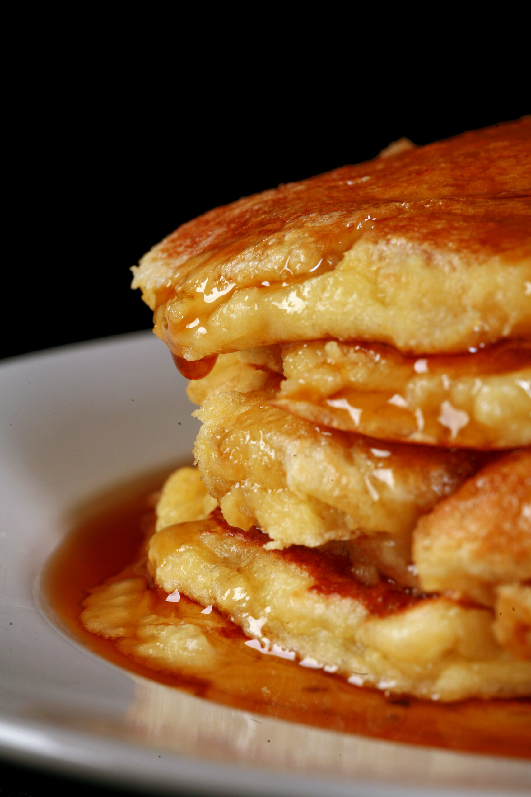 A stack of 6 banana protein pancakes on a place, with maple syrup tripping off the top.