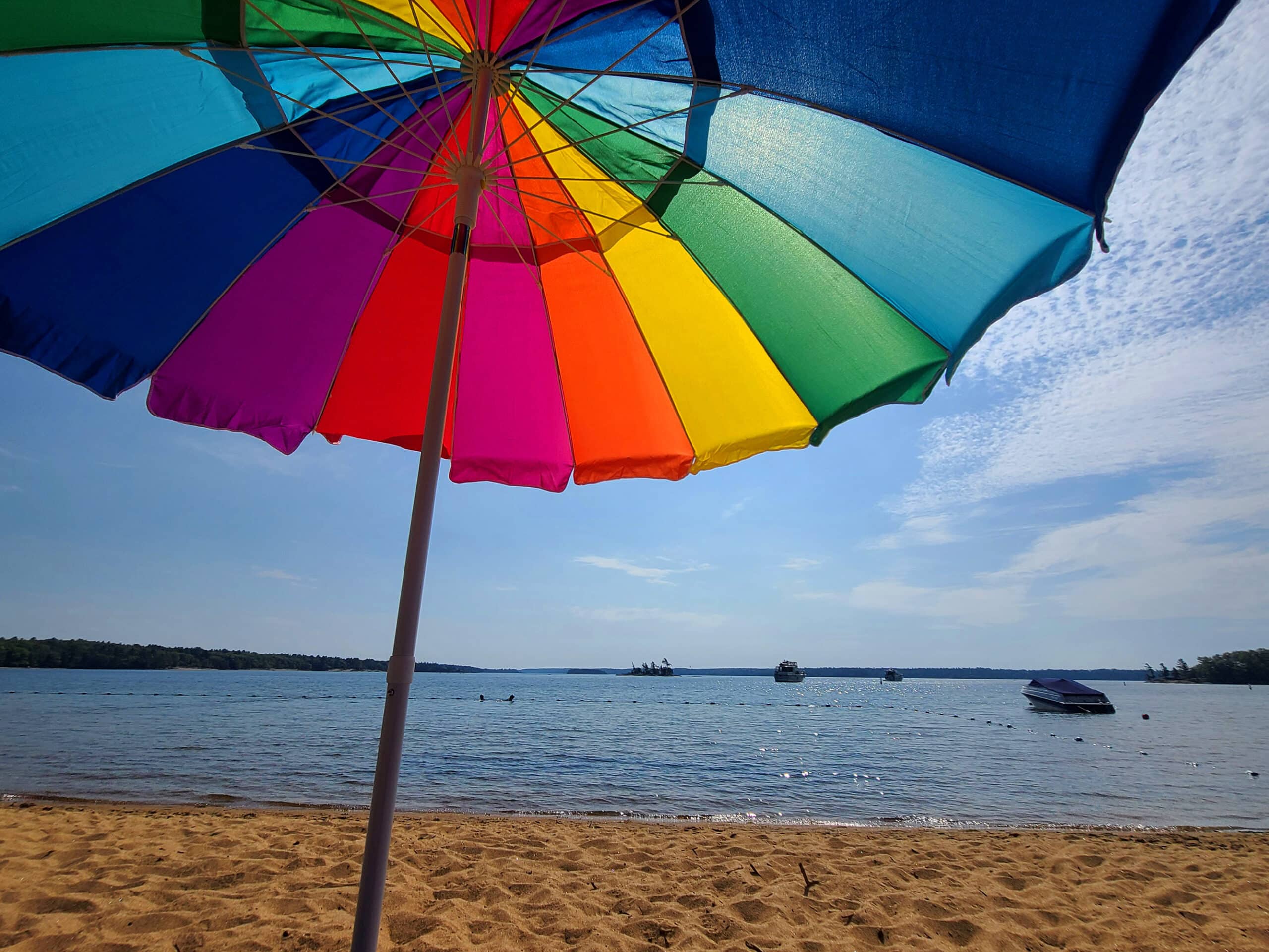 The beach and lake at Kilcoursie Bay.  A rainbow coloured beach umbrella is in the foreground.
