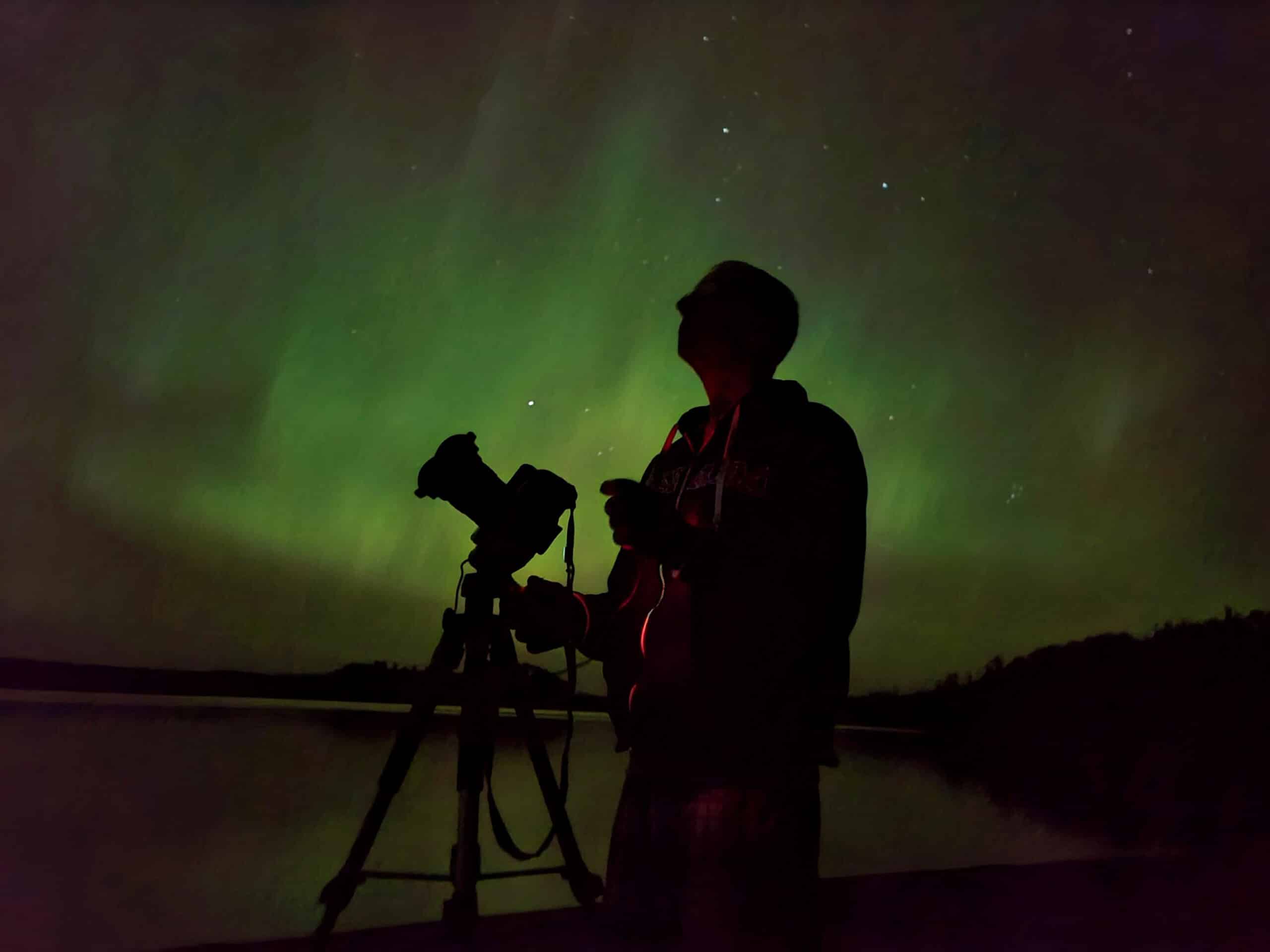 The silhouette of a man with a camera, with the aurora borealis behind him.