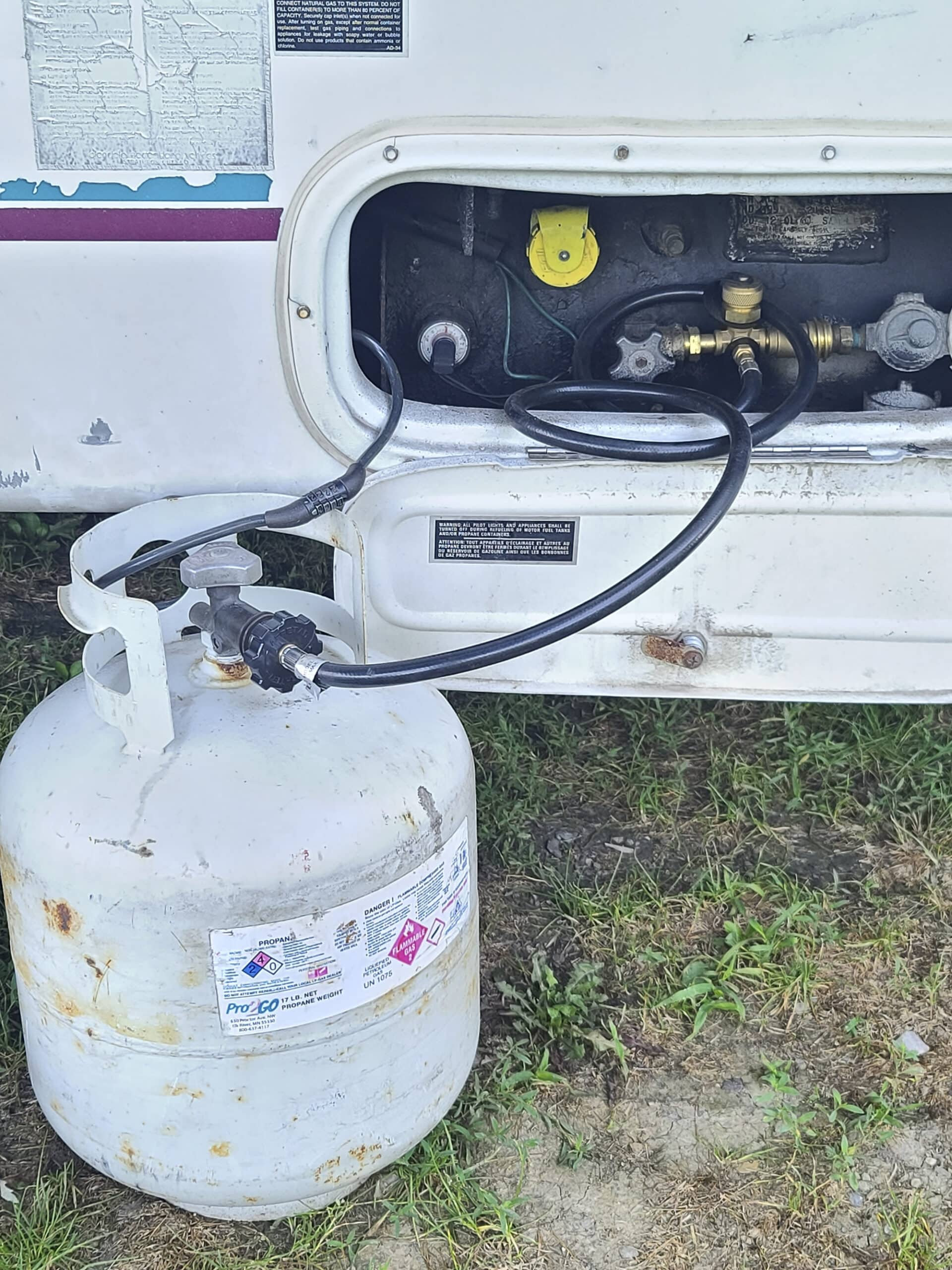 The propane tank and fill port of an RV. A barbecue propane tank is seen connected via a black hose.