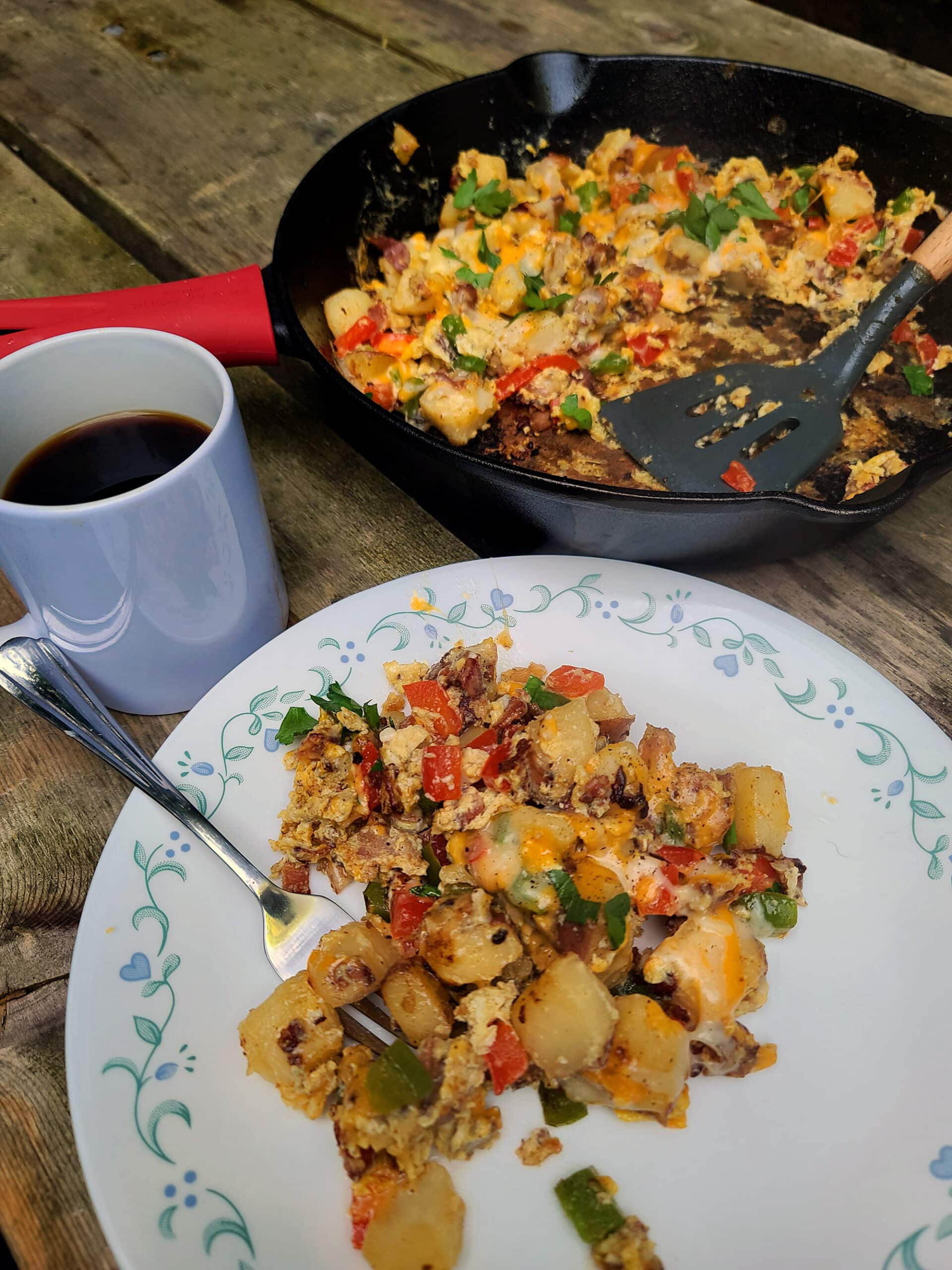 A plate of camping breakfast skillet in front of the cast iron pan, all laid out on a wooden picnic table.