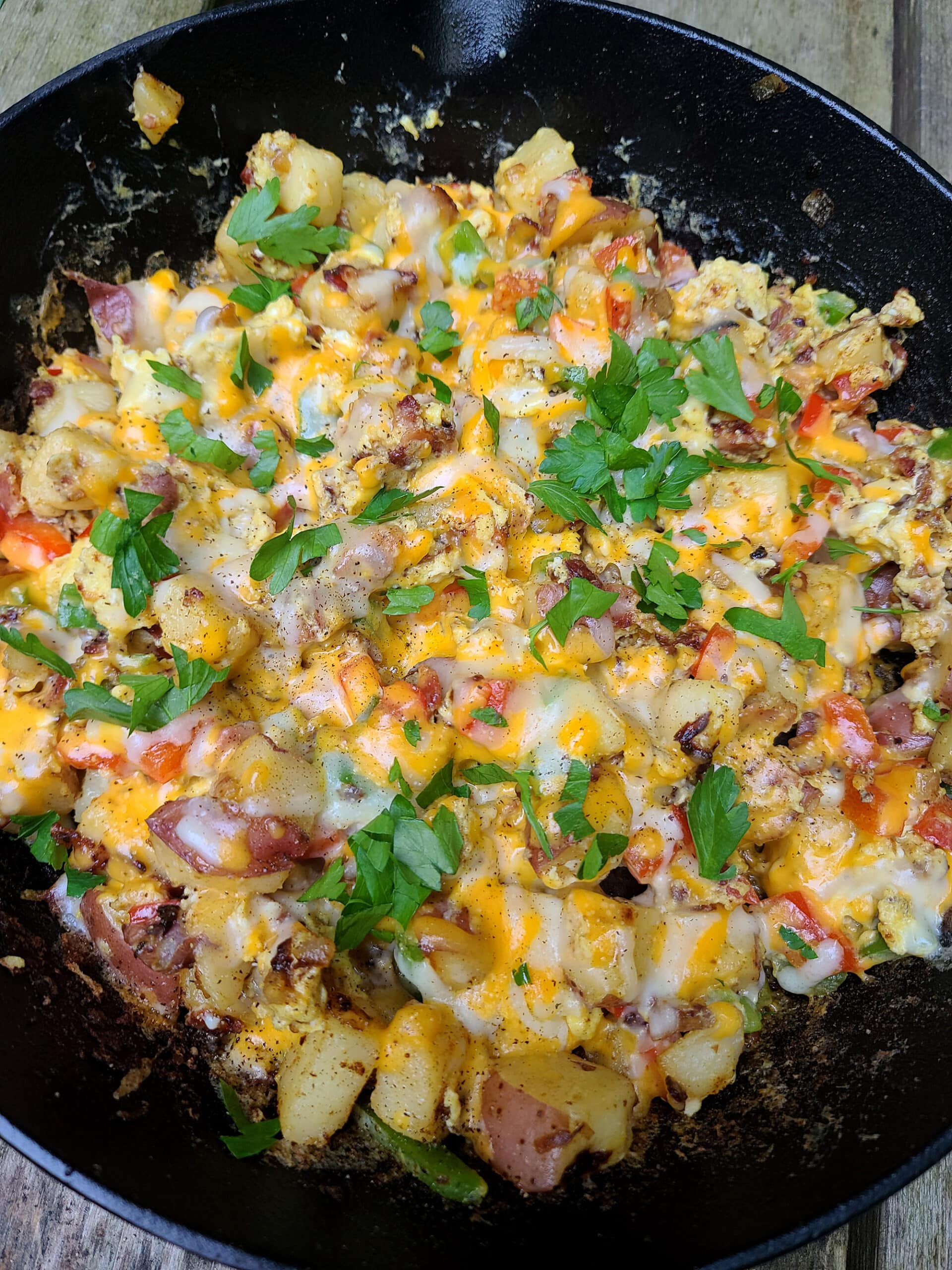 A breakfast skillet of potatoes, peppers, bacon, eggs, and cheese in a cast iron skillet, topped with parsley.