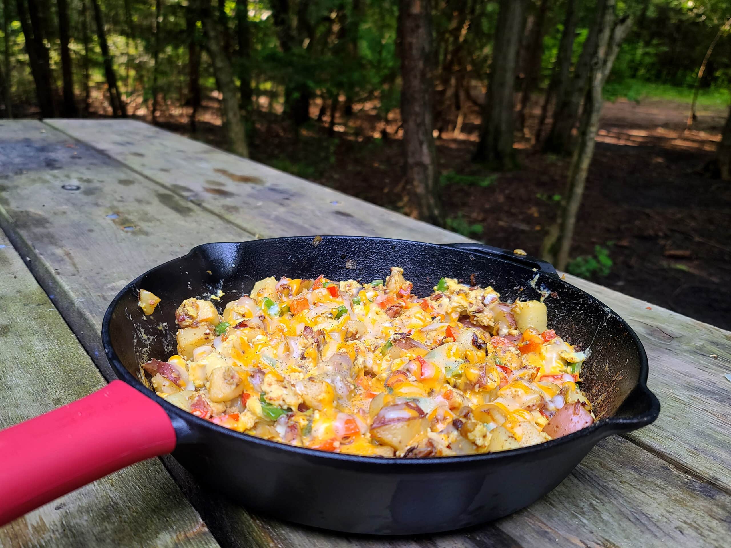 A cast iron pan with potatoes, eggs, peppers, cheese, and bacon, resting on a camping table.