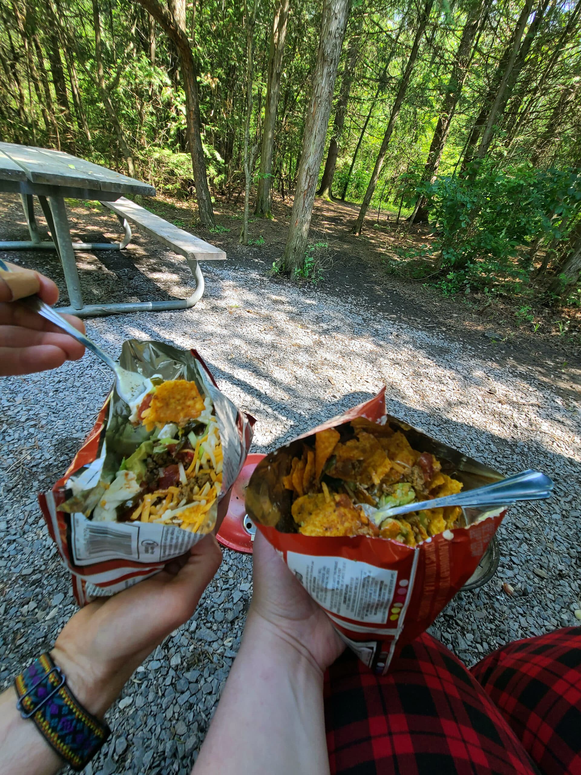 Two people sitting by a camp site, holding walking taco bags in front of them.
