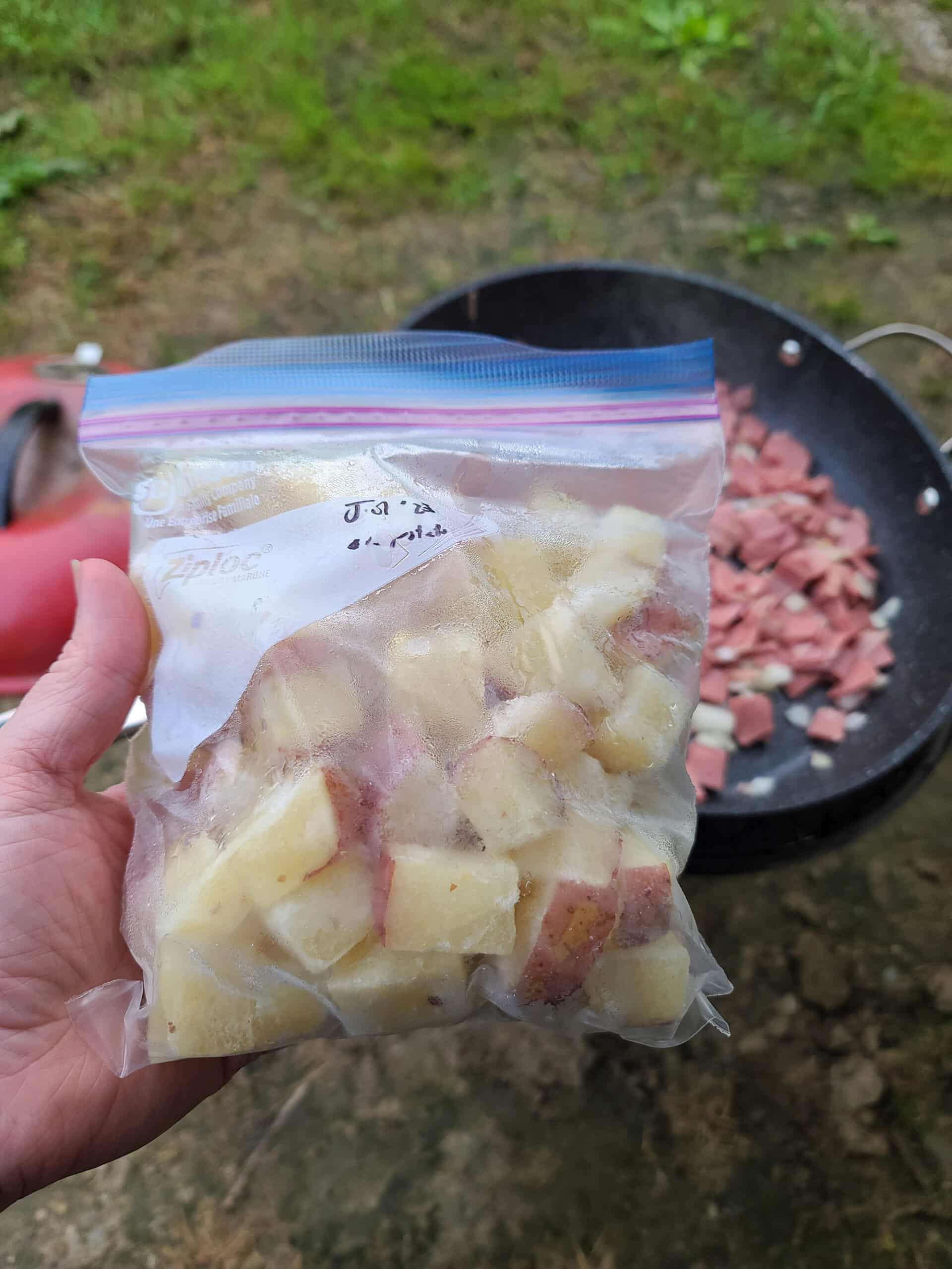 A hand holds a bag of frozen potato cubes in front of a skillet over a charcoal grill.