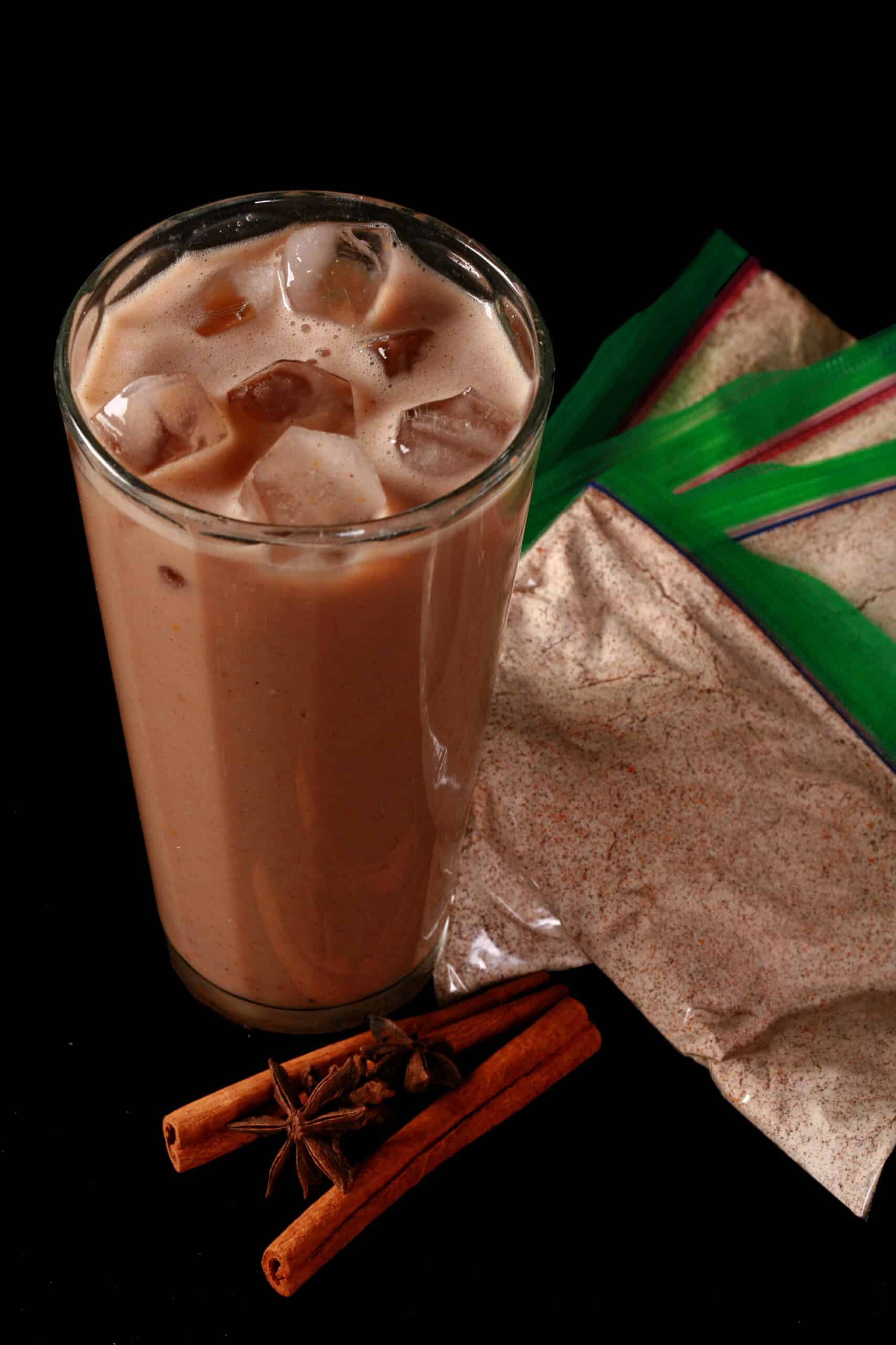 A tall glass of protein iced pumpkin chai latte next to several baggies of pumpkin chai latte mix.