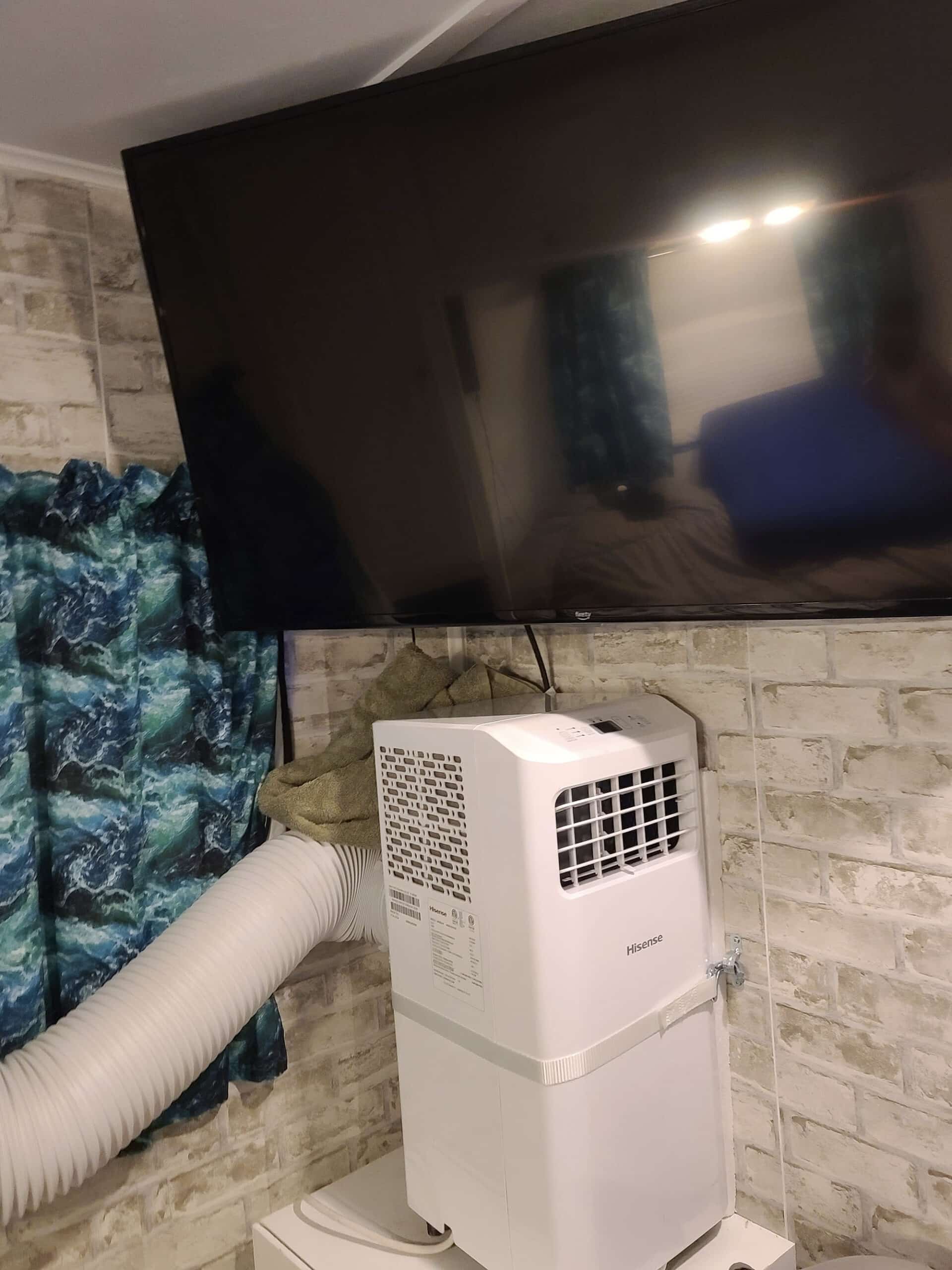 A large TV is off, mounted on the wall.  Underneath is a white portable air conditioner, mounted against the wall.