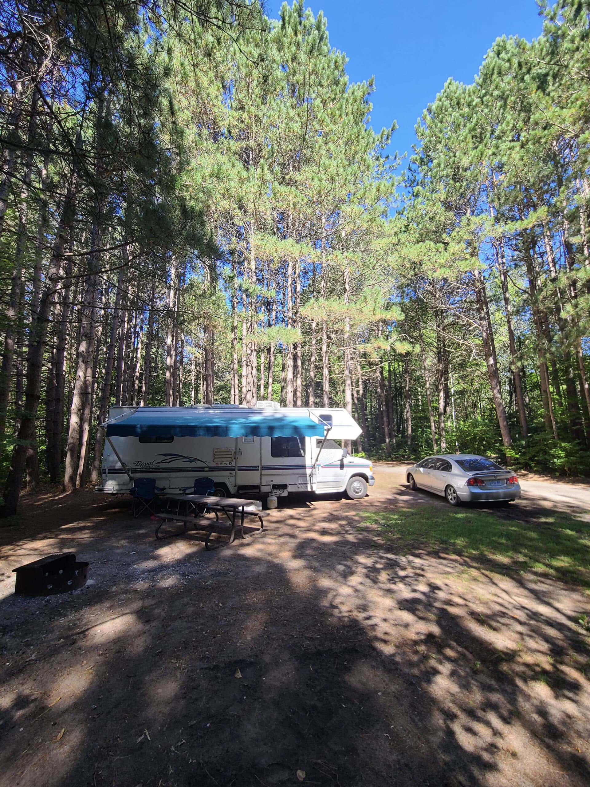 An RV and car parked at a camp site.  Tall trees block much of the light on the site, though it is a sunny day.