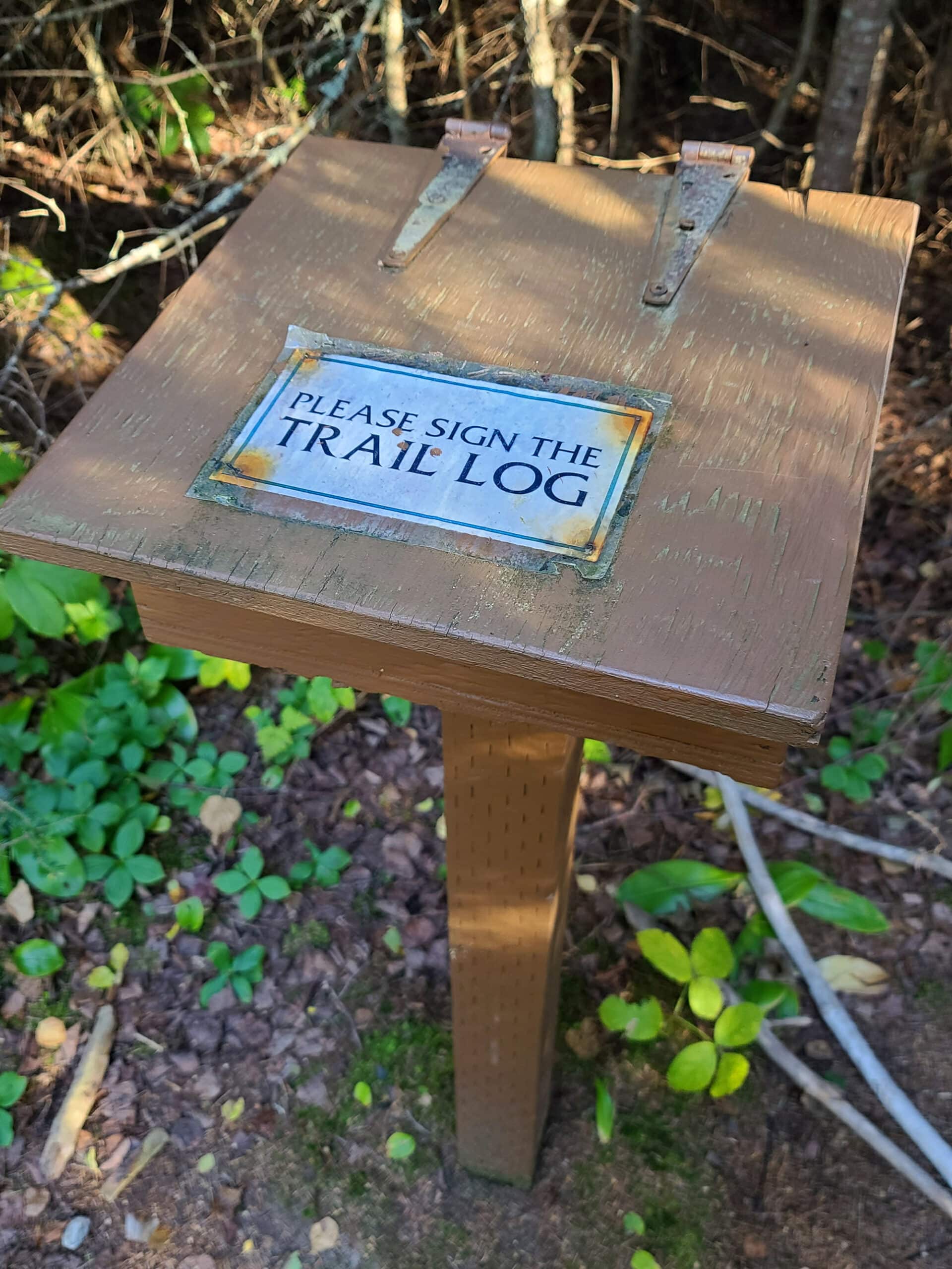 A wooden pedestal with a label asking hikers to please sign the trail log.