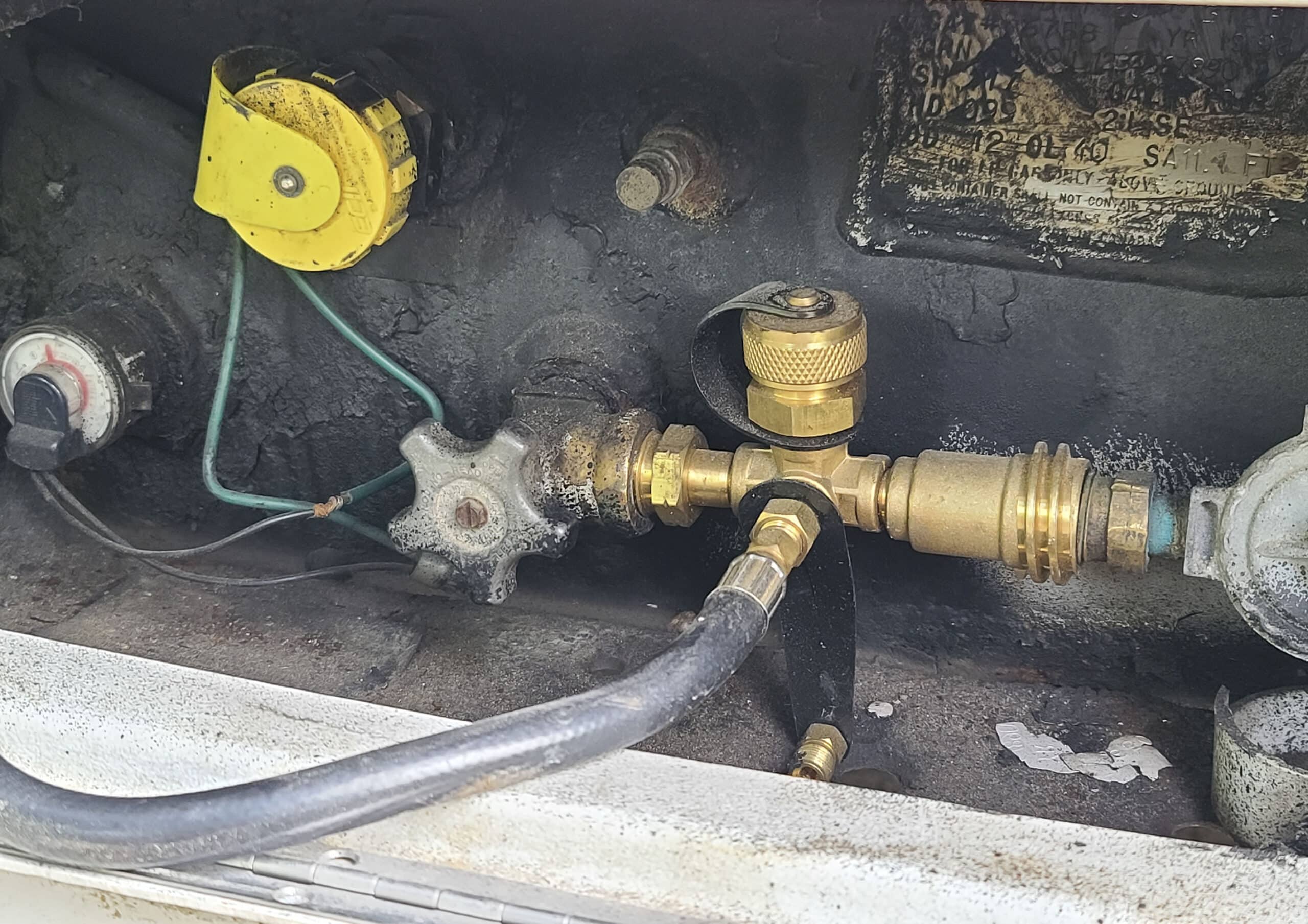 A propane port and valve on a motorhome.  A brass tee has a black rubber hose leading away.