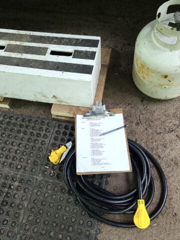 An RV electrical cord coiled on the ground. On top is a clipboard with a checklist and a pen. In the background is a rubber mat and wooden step.