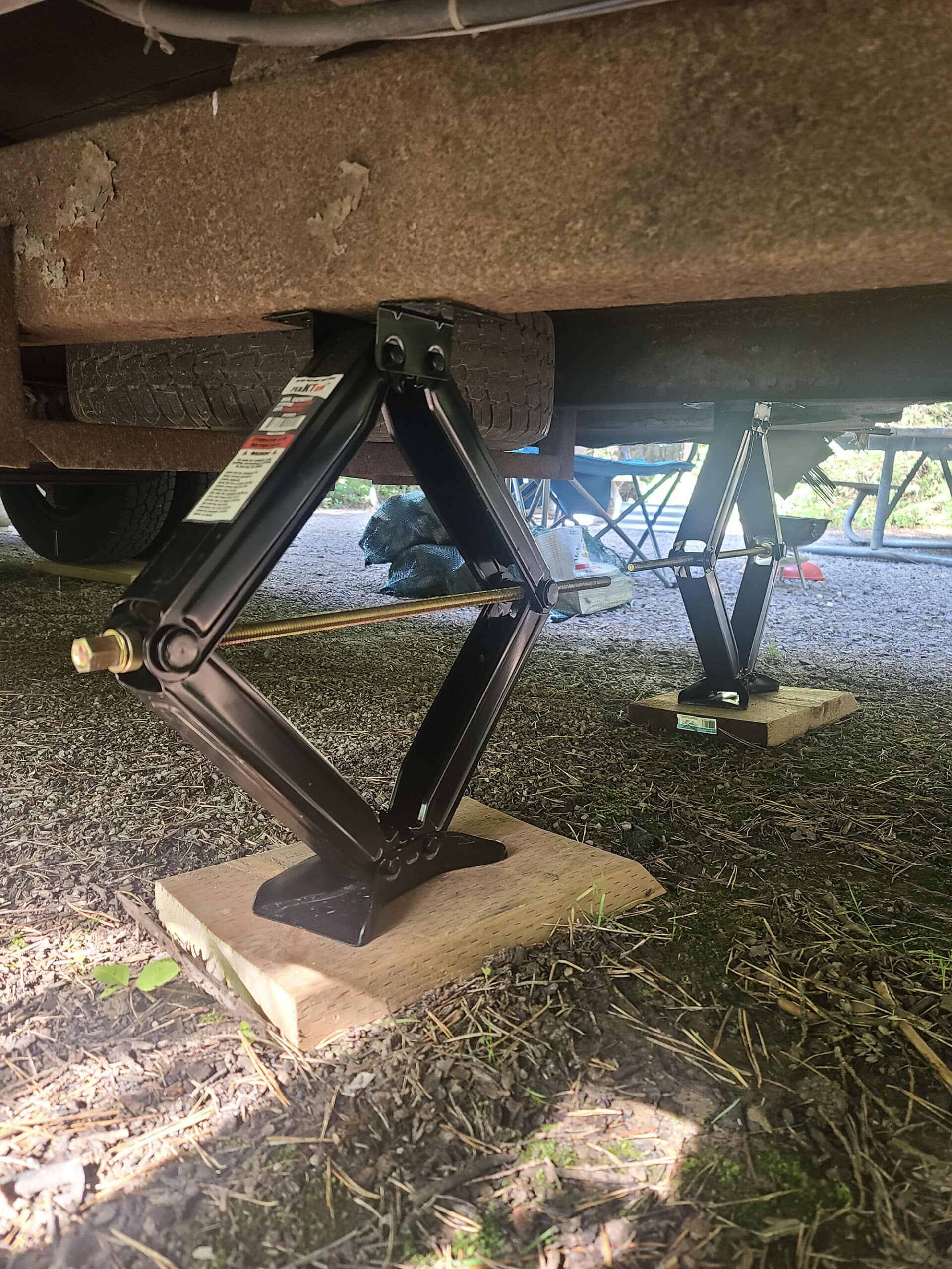 A low view underneath an RV with two scissor jacks, one supporting each frame rails.  There is a a piece of wood under each jack.