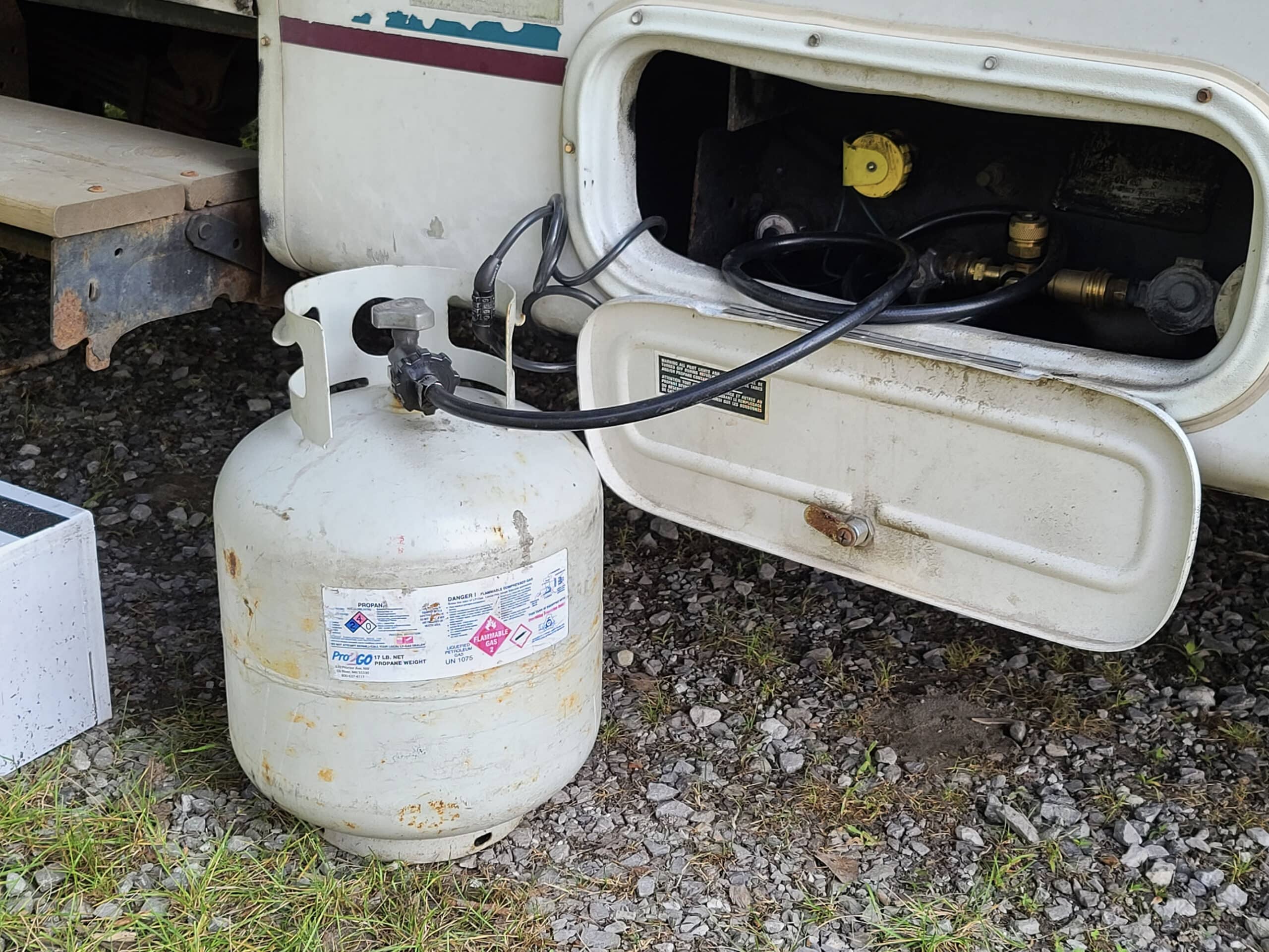 A white barbeque propane tank connected to a motorhome with a black hose.
