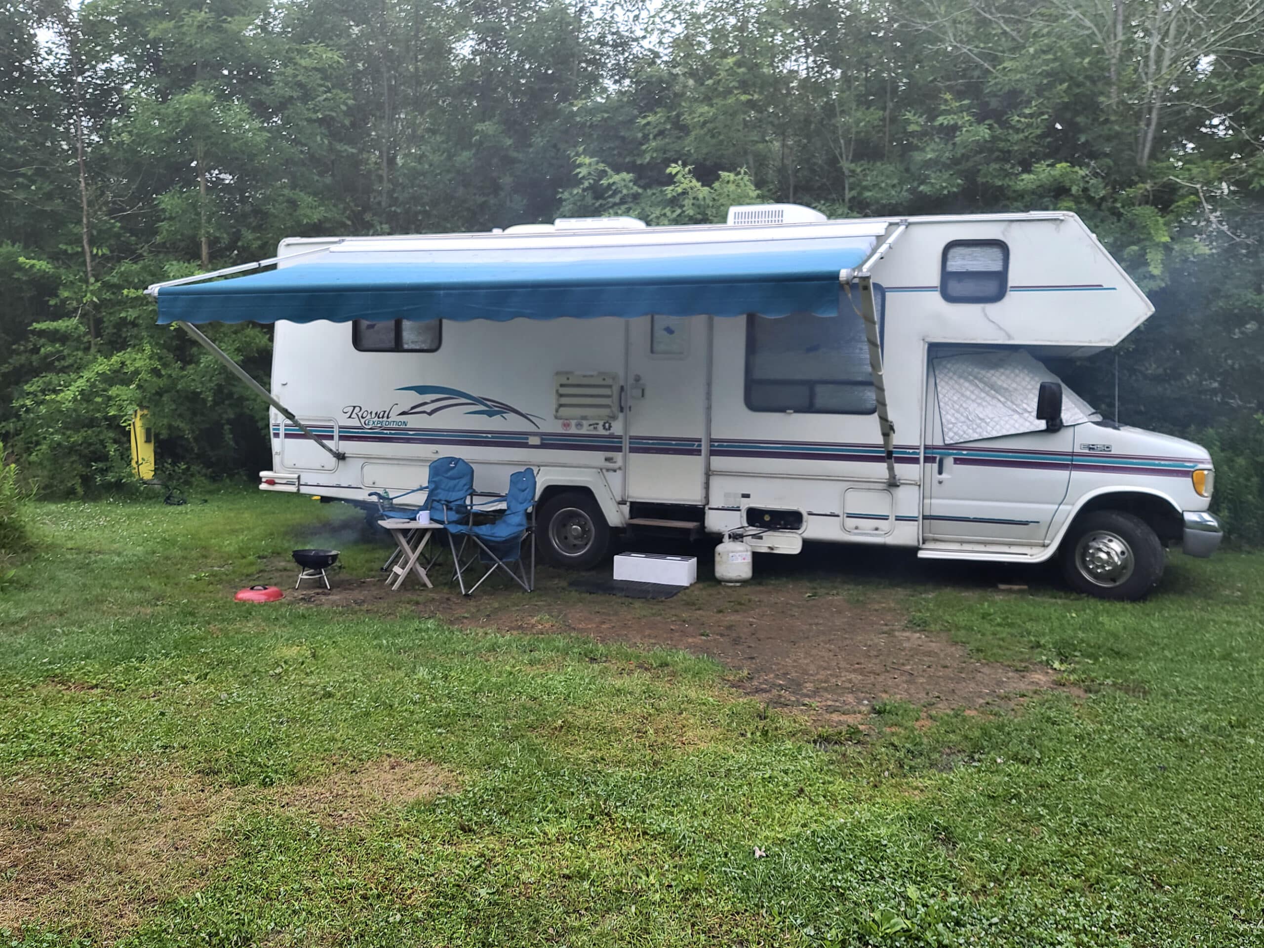 A side view of a motorhome setup at a camp site with awning out, two chairs, and a small grill.