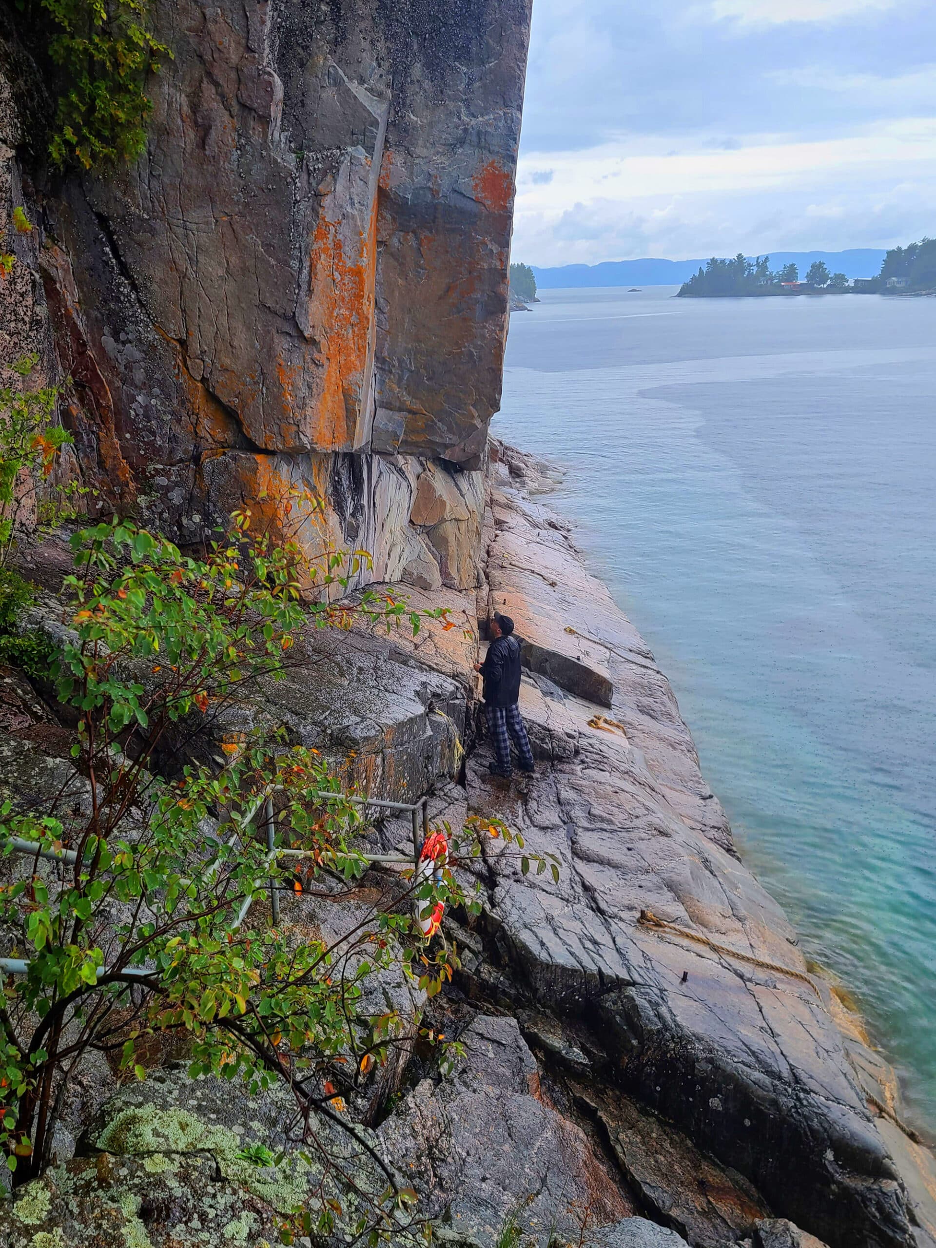 A long view photo looking down at a man standing on rocks, lake superior behind him.  He is looking at the Agawa Bay Rock Pictographs,