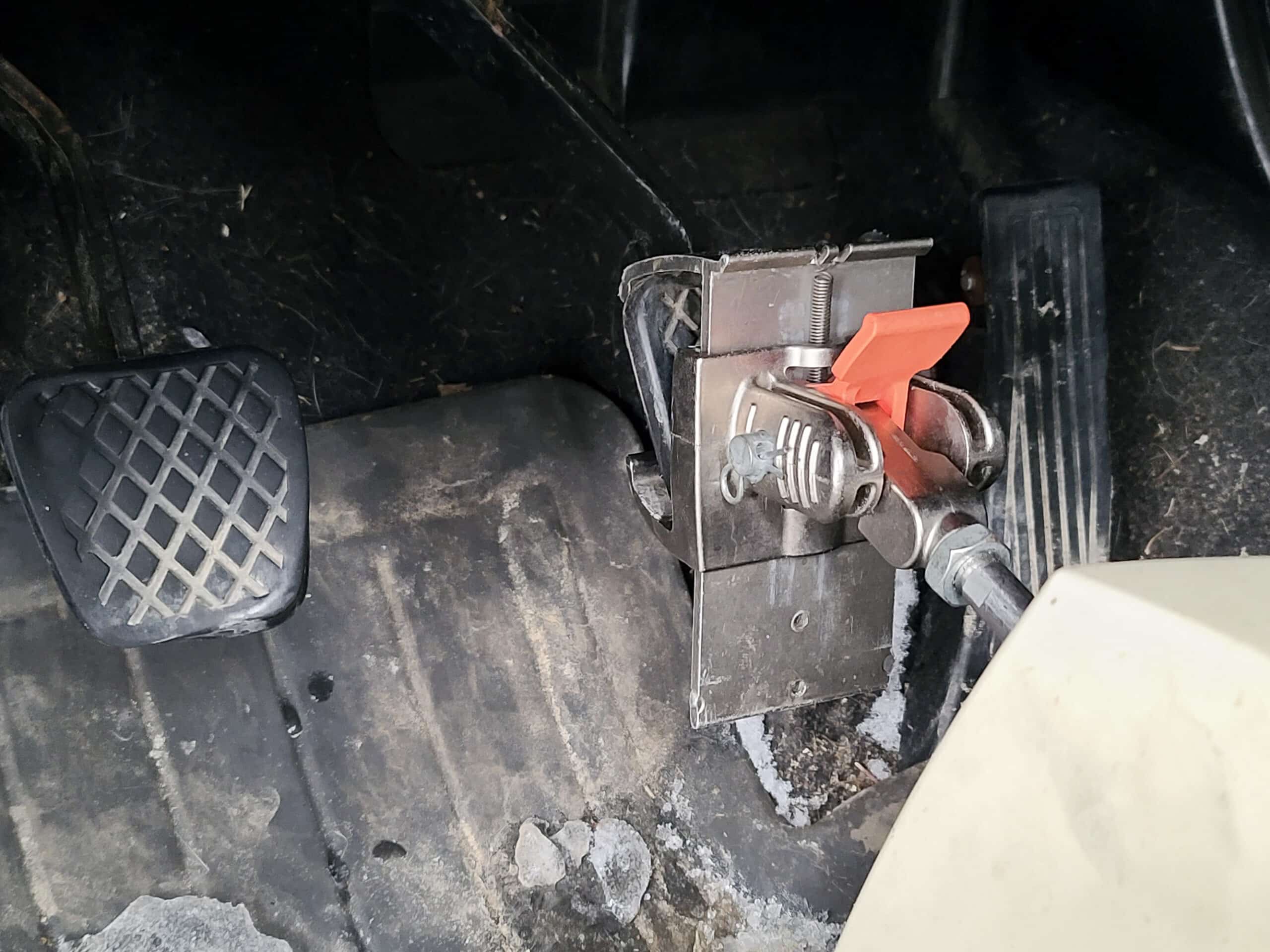 A metal bracket attached to the brake pedal of a car.  A rod connects the clamping mechanism to a white container.