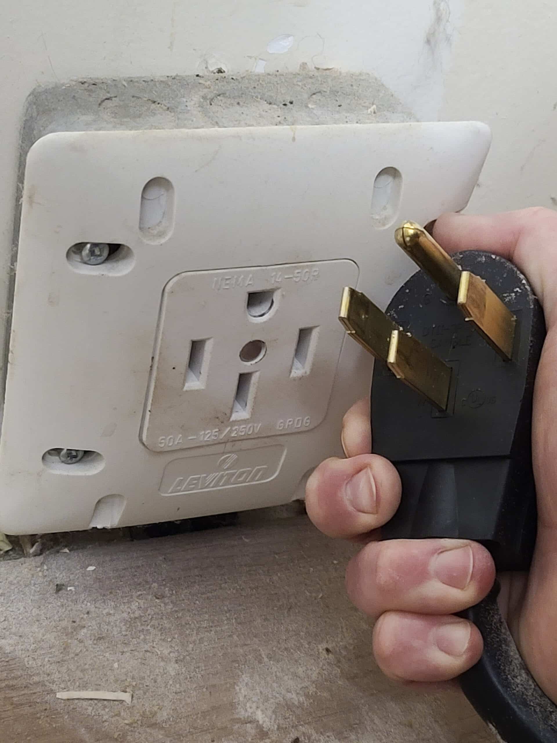 a 120/240v plug held next to a matching outlet mounted on a wall, near the floor.