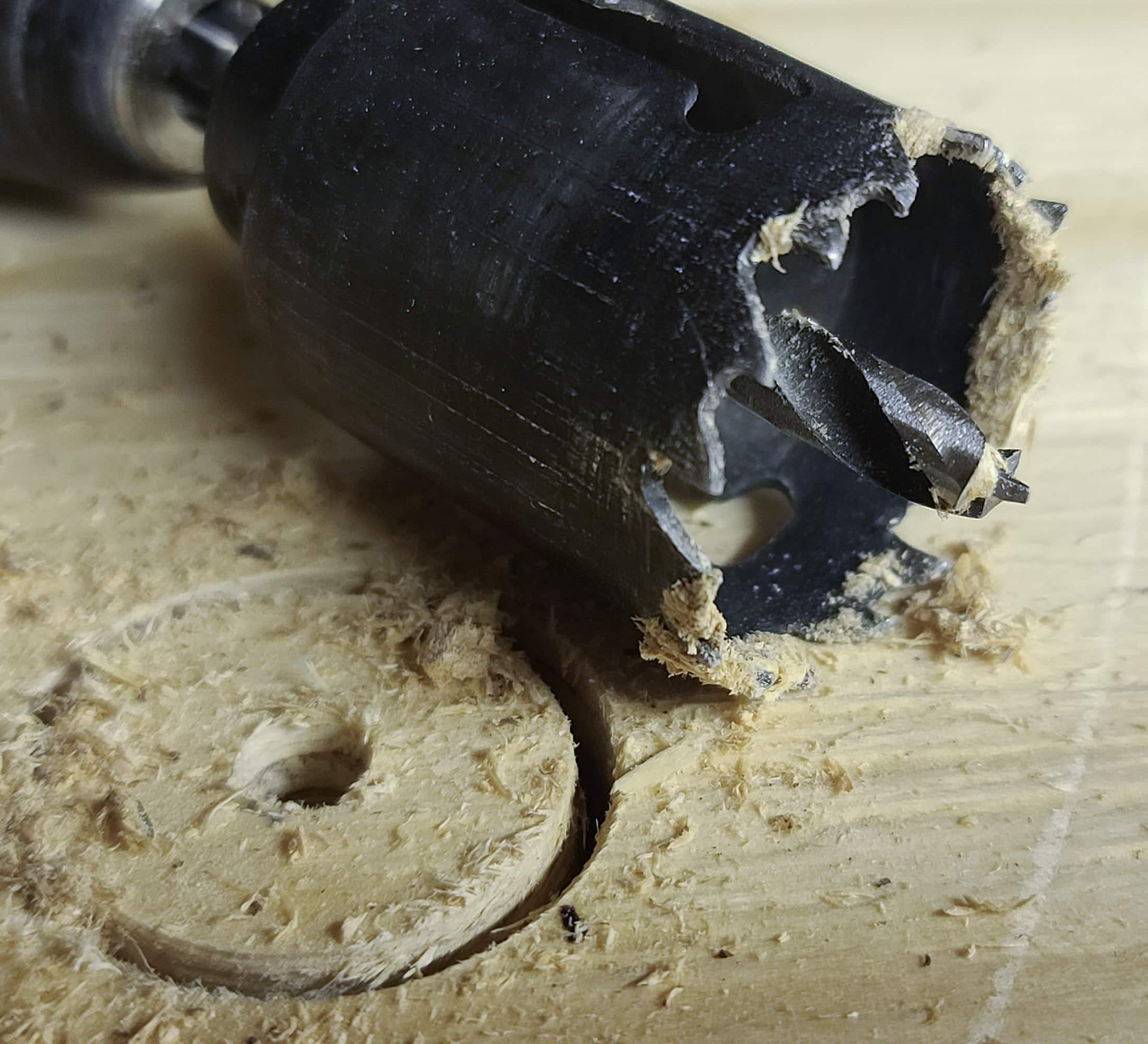 A closeup of a hole saw on a board. The hole saw has sawdust stuck to the teeth. The board has a hole partially cut out.