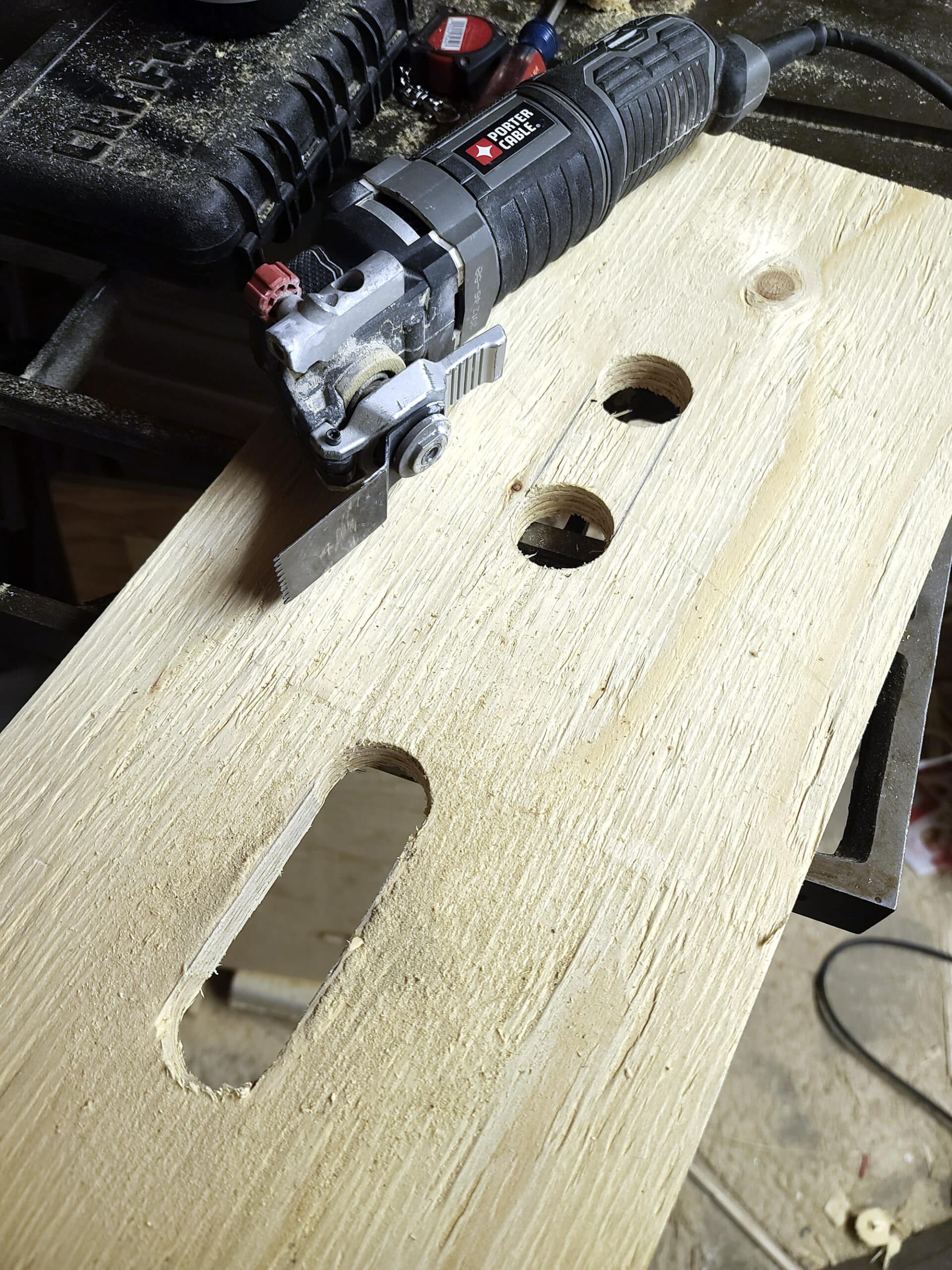 A wood board with two holes near each other, and an oval cutout on the other side of the board. An oscillating cutting tool is on top.