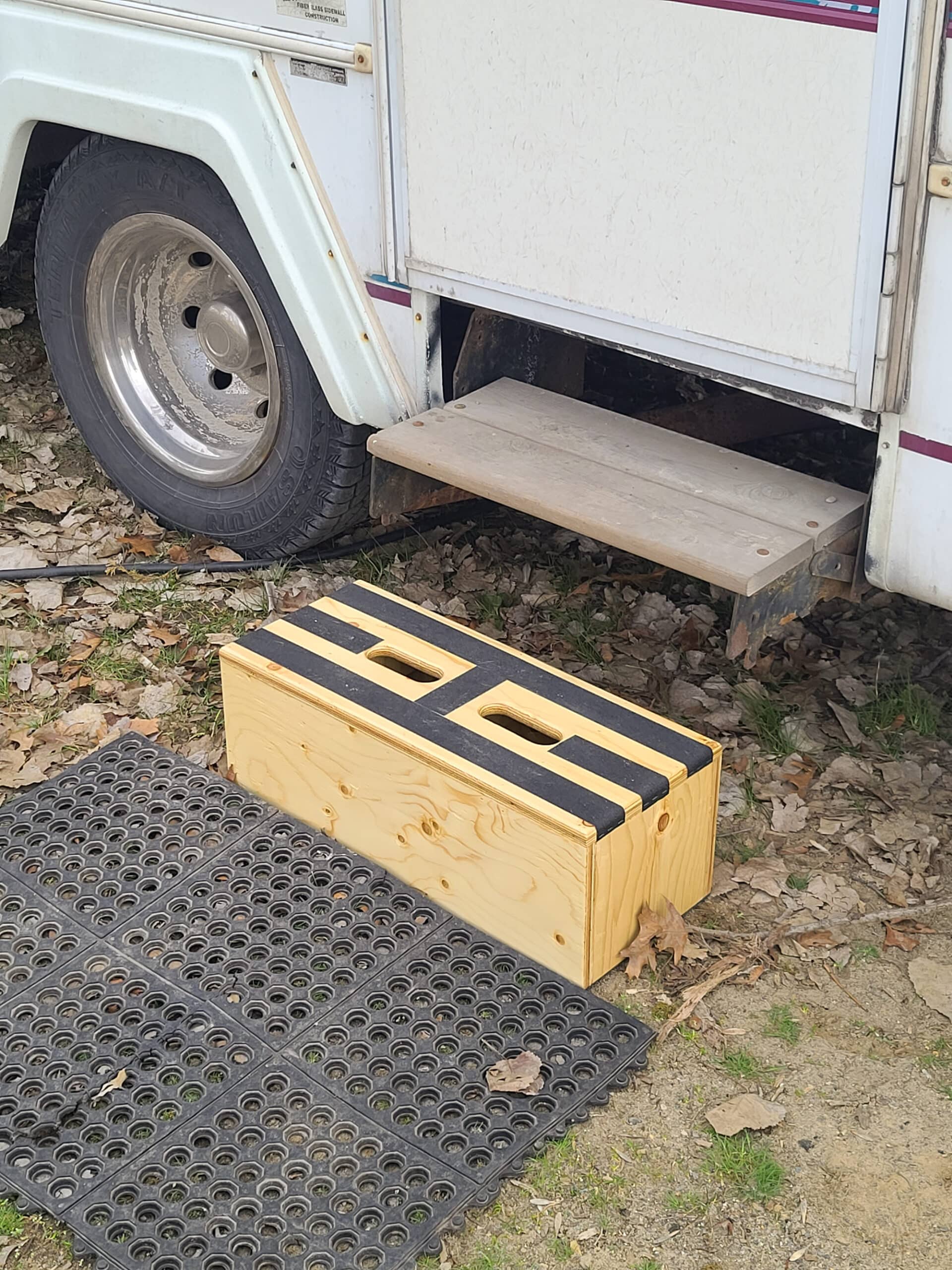 A motorhome with a single fold out step very high. A wood step below it, and a rubber mat on the ground. Overlaid text says Build Your Own RV Camper Step.