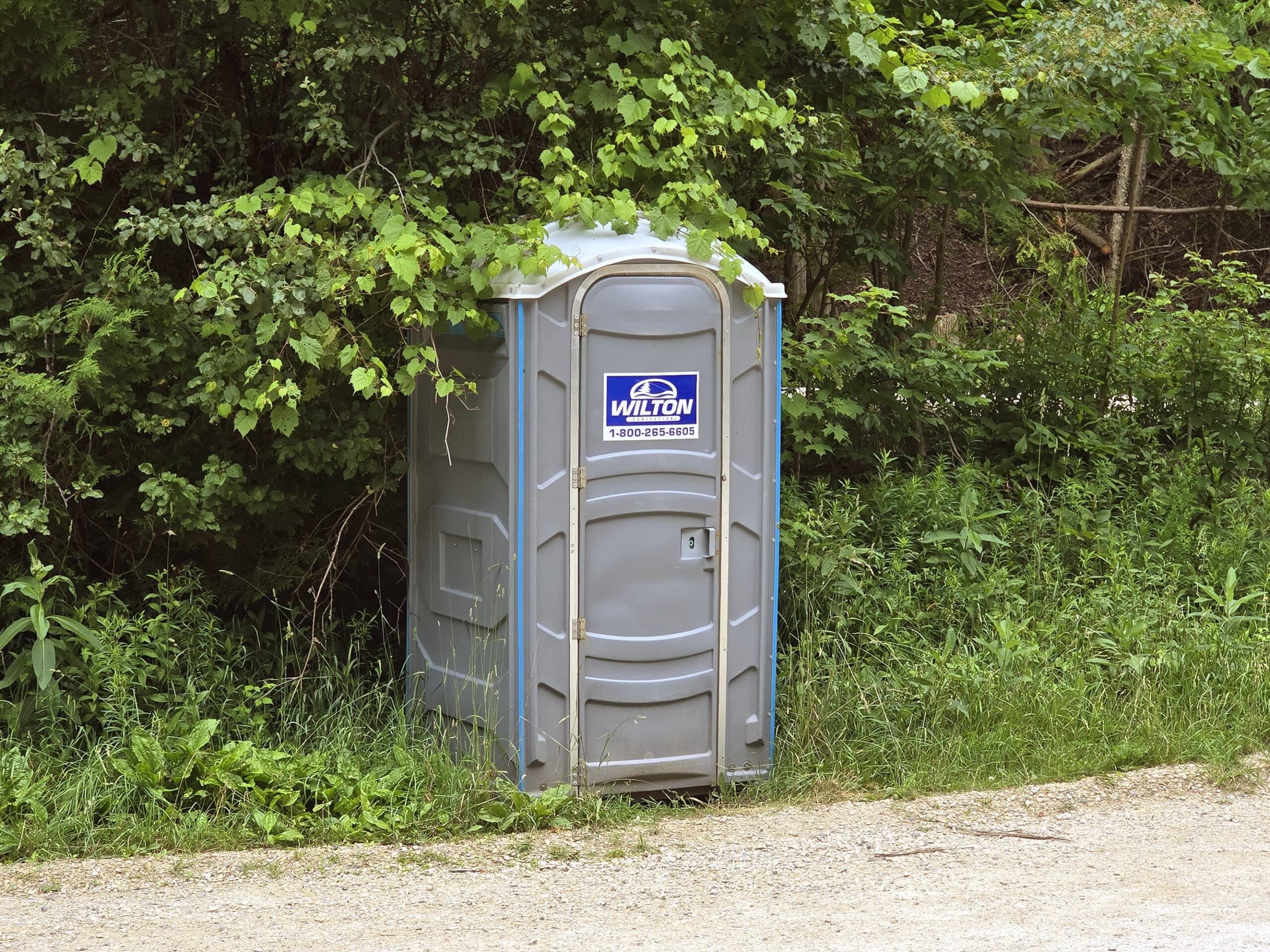 A grey portapotty on the side of the parking lot.