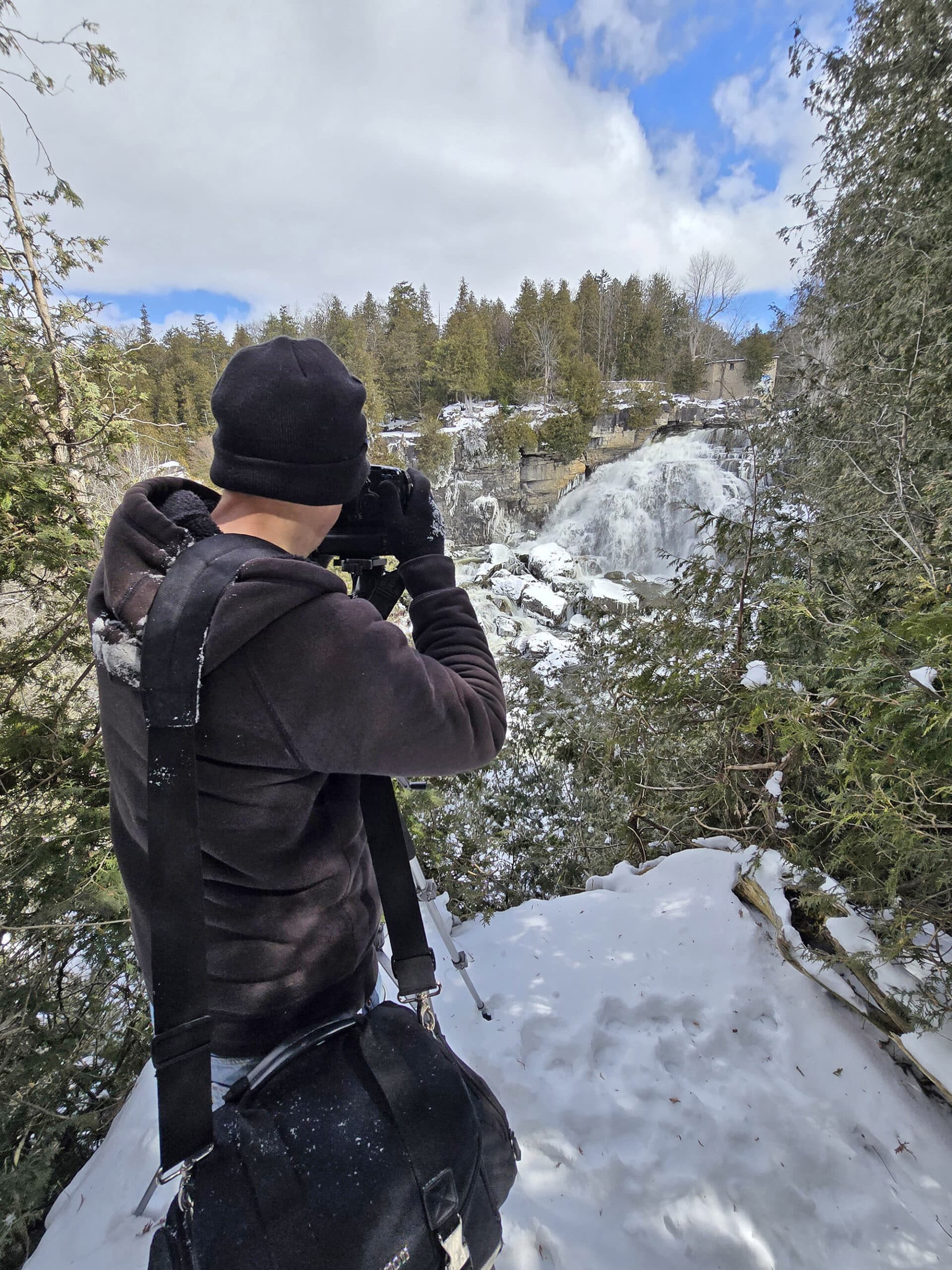 A middle aged white man taking a photo of Inglis Falls in the winter.