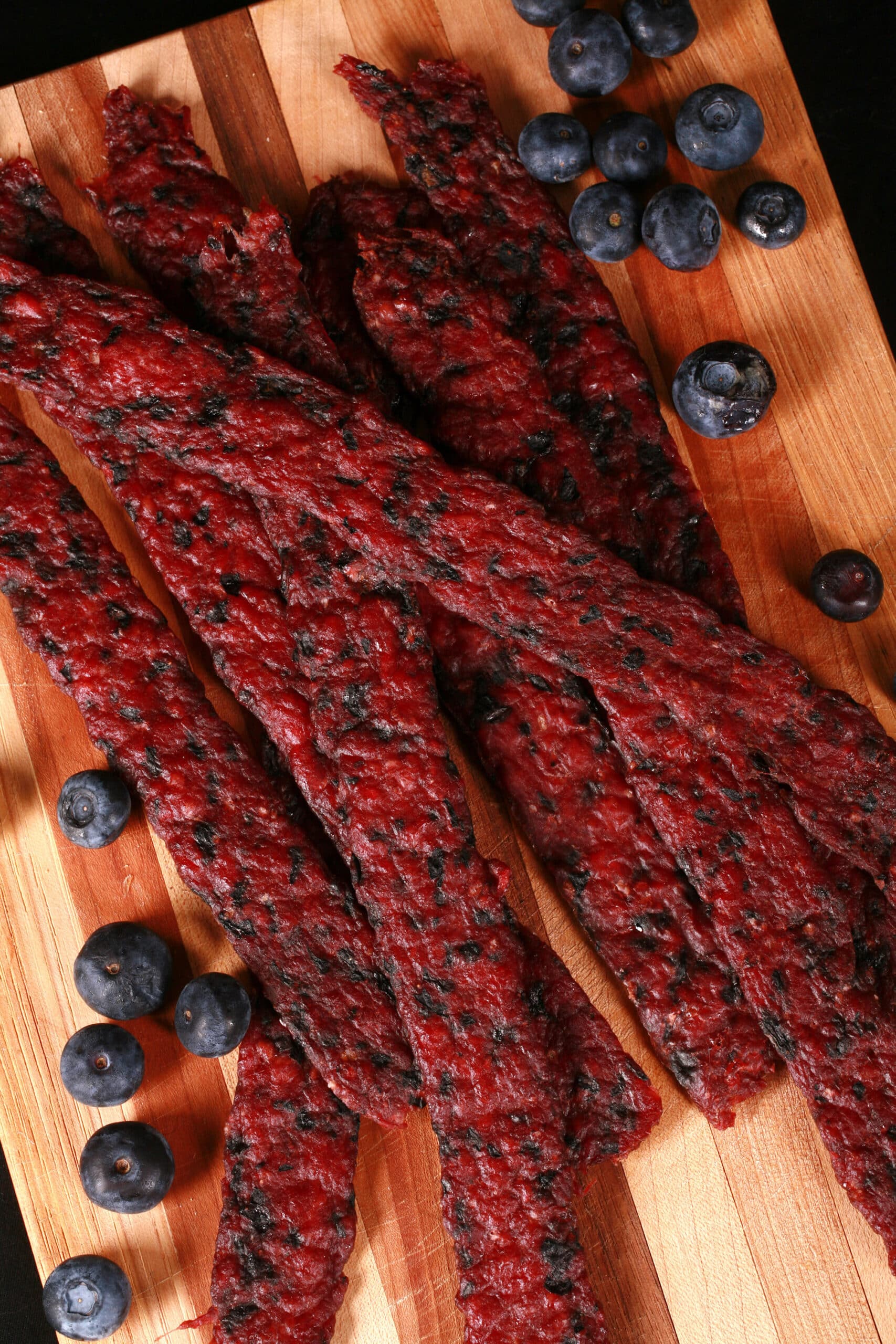 Several blueberry bison jerky strips on a cutting board with fresh blueberries.