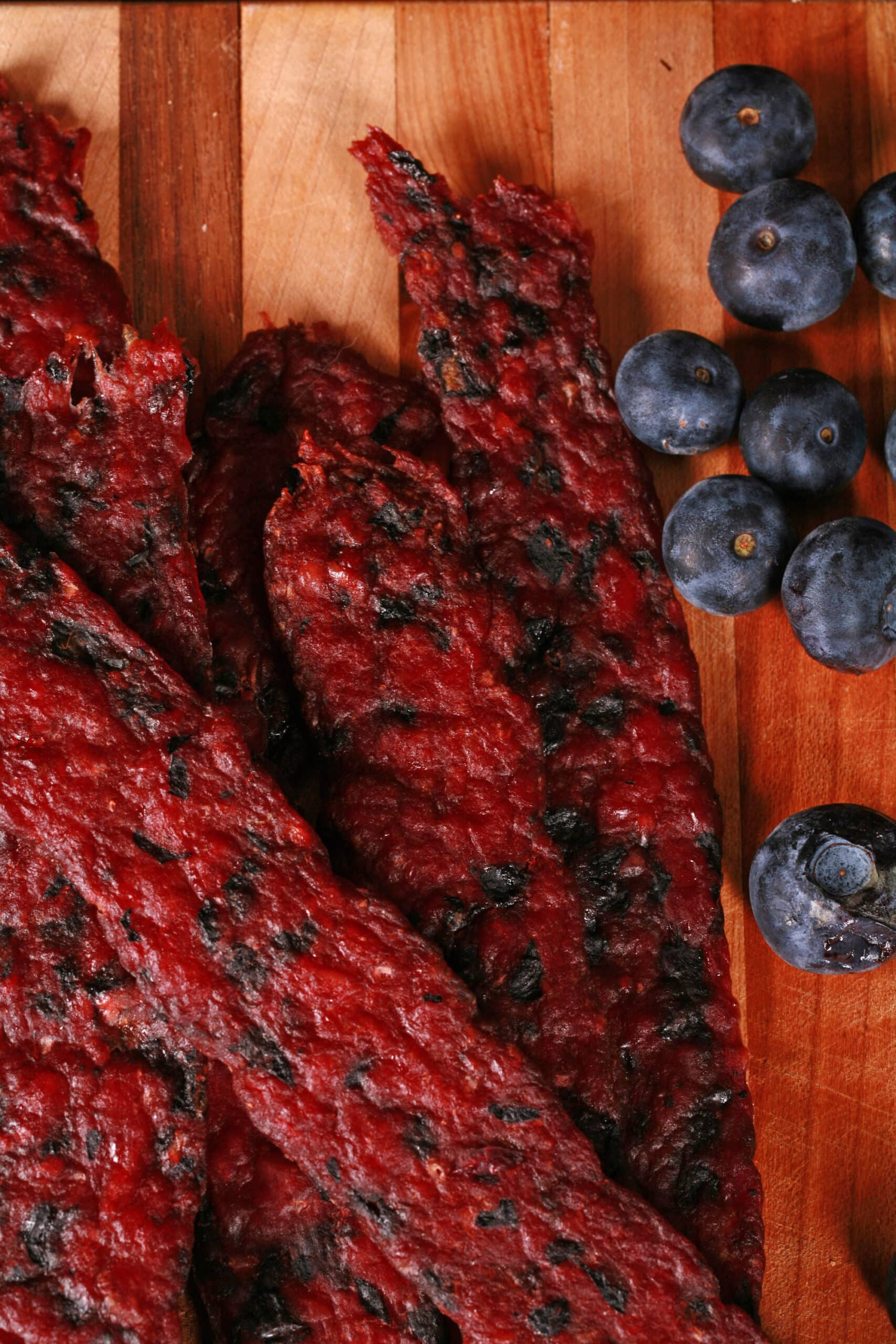 Several blueberry bison jerky strips on a cutting board with fresh blueberries.