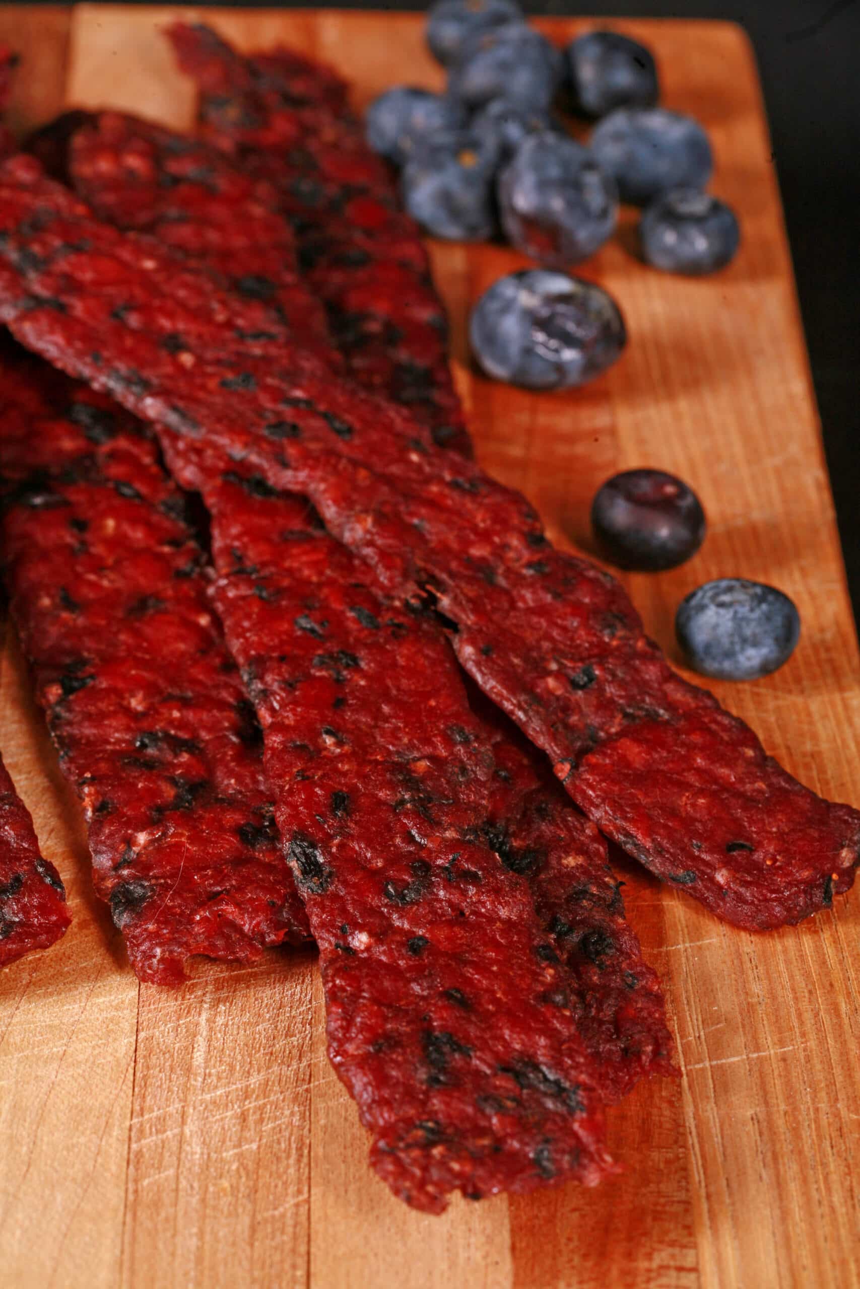 Several bison jerky strips on a cutting board with fresh blueberries.