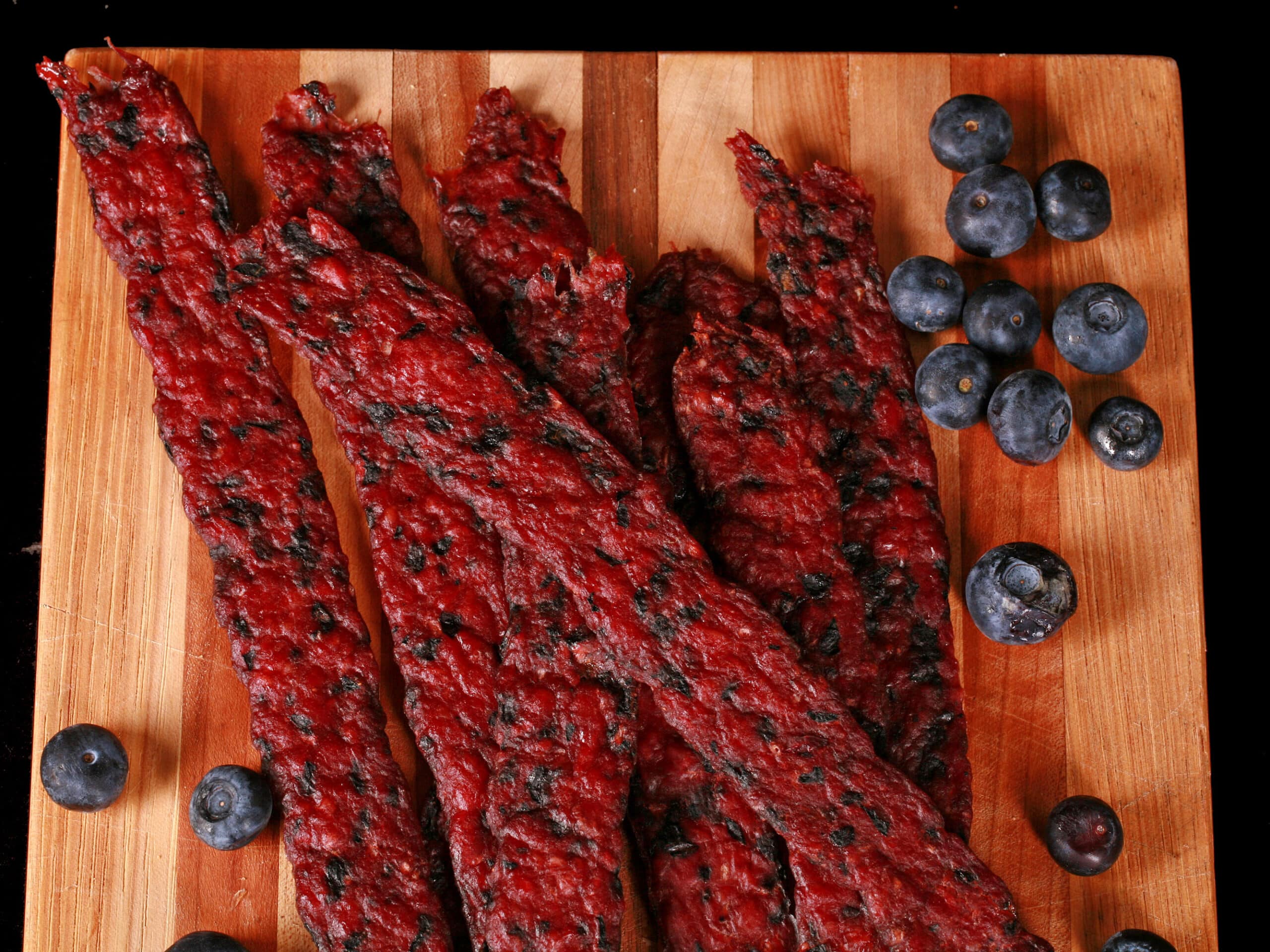 Several blueberry bison jerky strips on a cutting board with fresh blueberries.