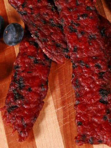 Several bison jerky strips on a cutting board with fresh blueberries.