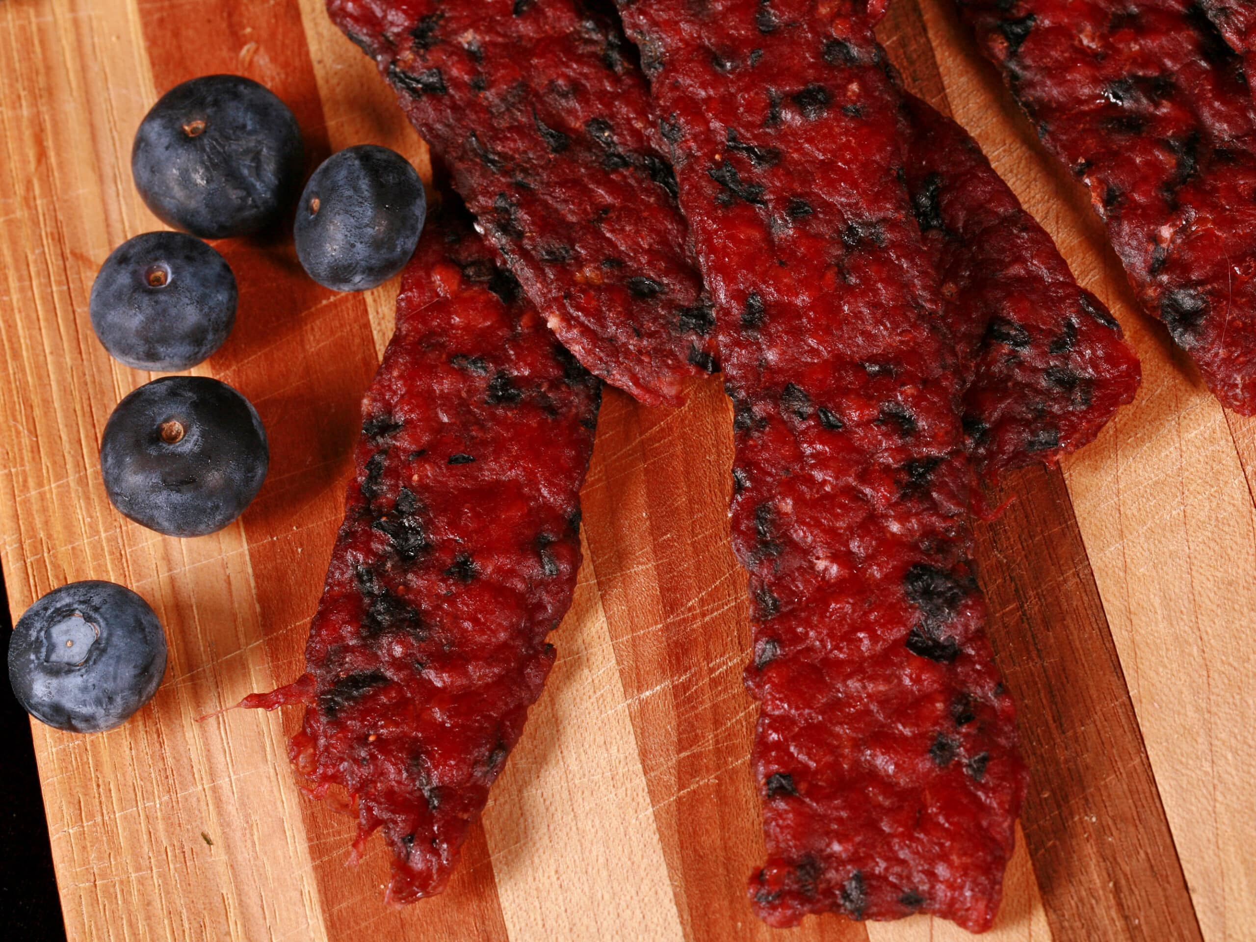 Several bison jerky strips on a cutting board with fresh blueberries.