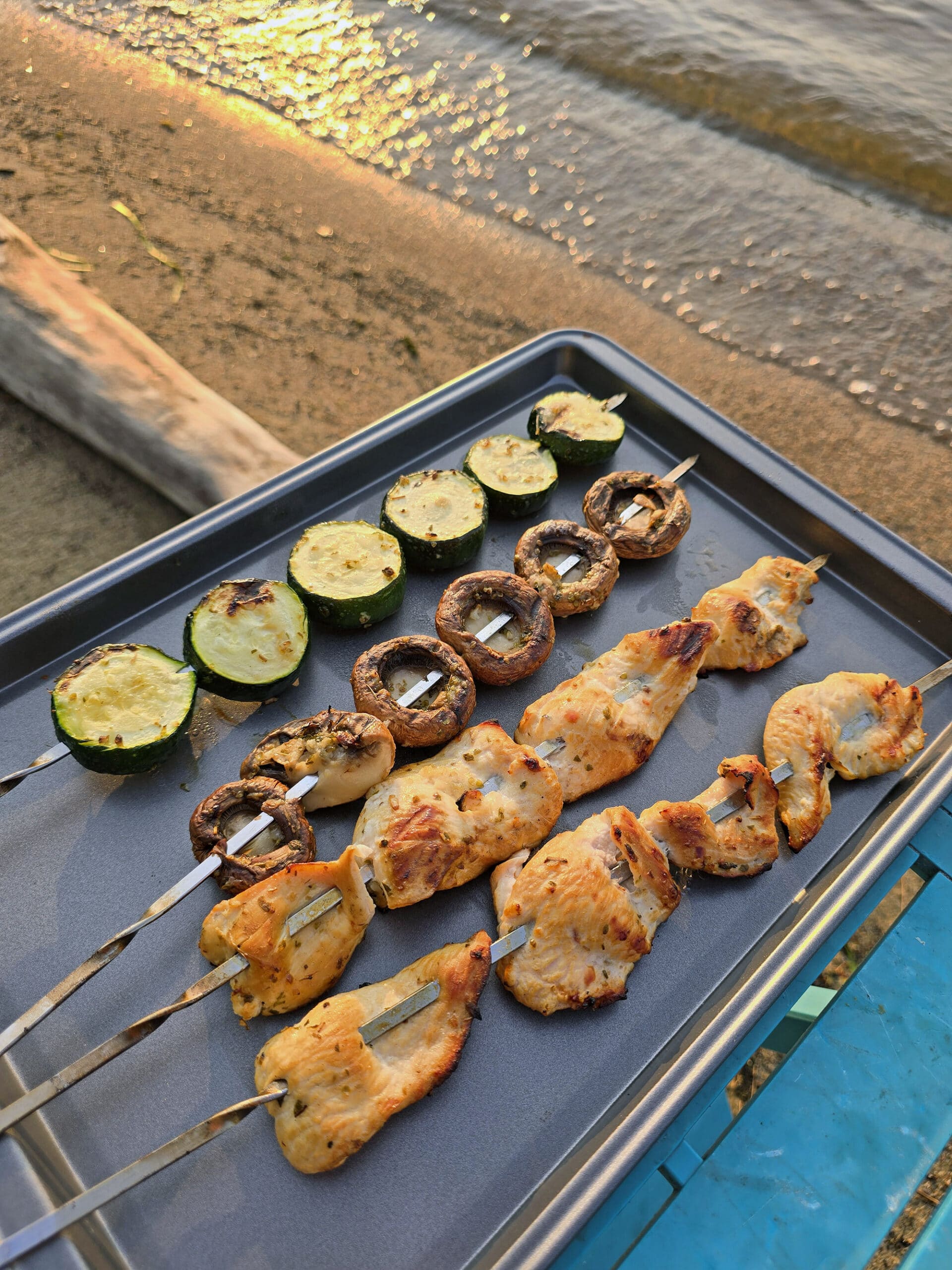 Mediterranean Chicken Kebabs  with skewers of zucchini and mushrooms on a tray with the beach in the background.