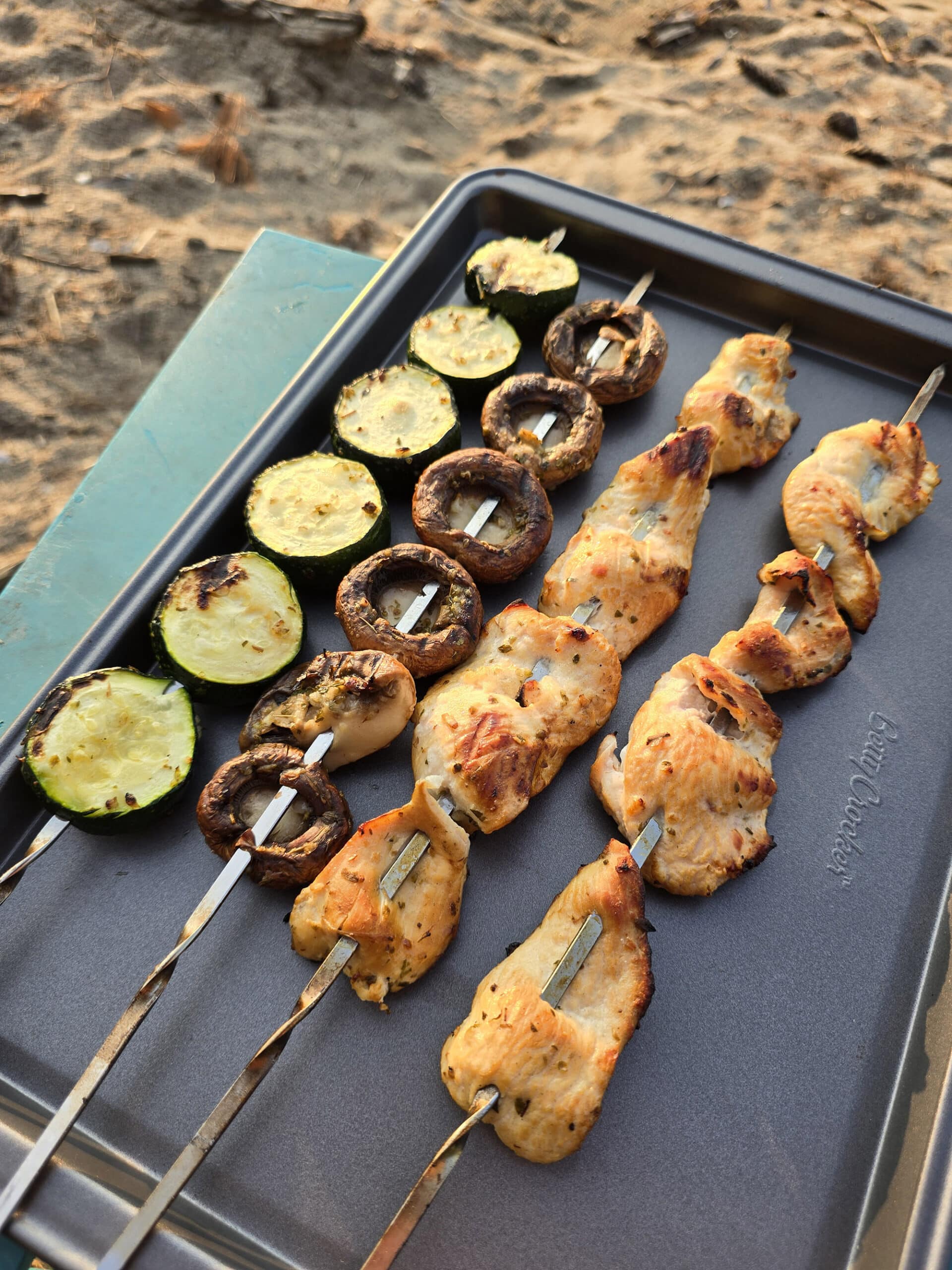 Mediterranean Chicken Kebabs  with skewers of zucchini and mushrooms on a tray with the beach in the background.