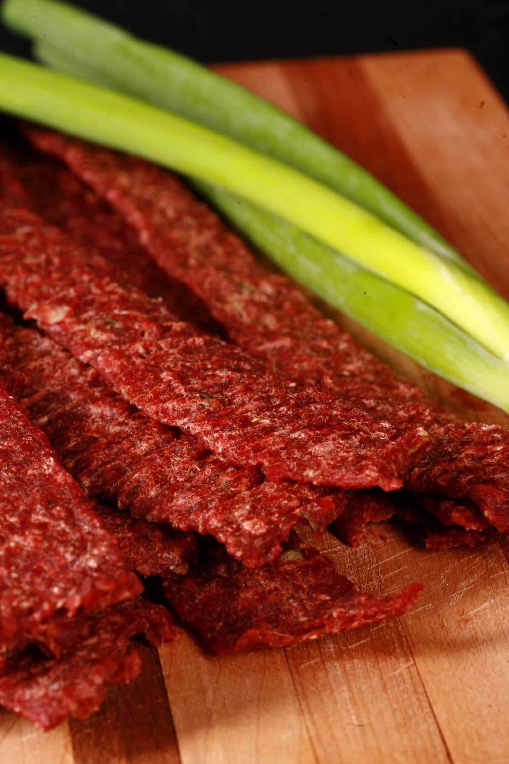 A pile of homemade ginger ground beef jerky on a wooden cutting board with green onions.