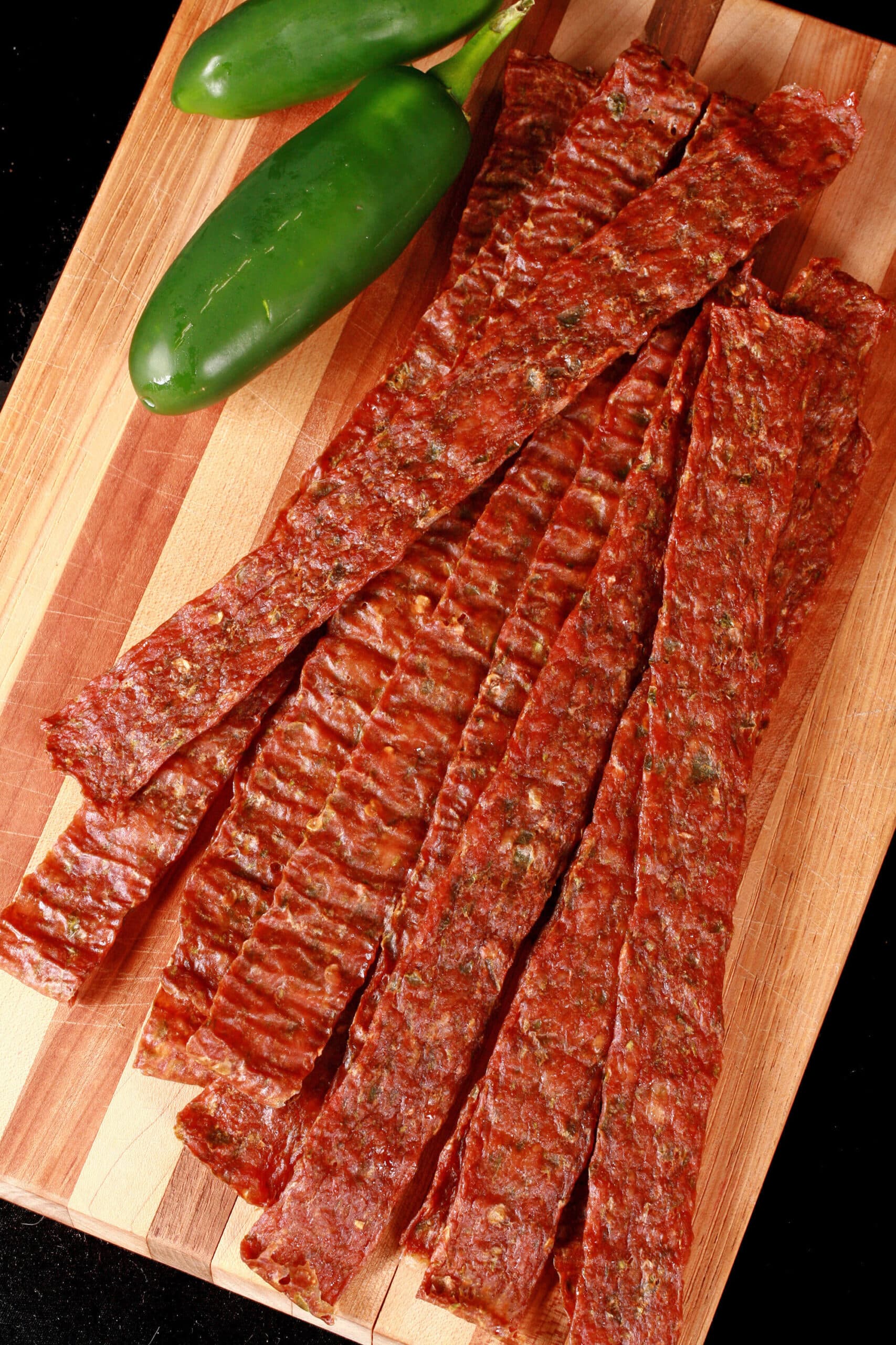 A pile of homemade chicken jerky on a wooden cutting board, along with 2 jalapenos.