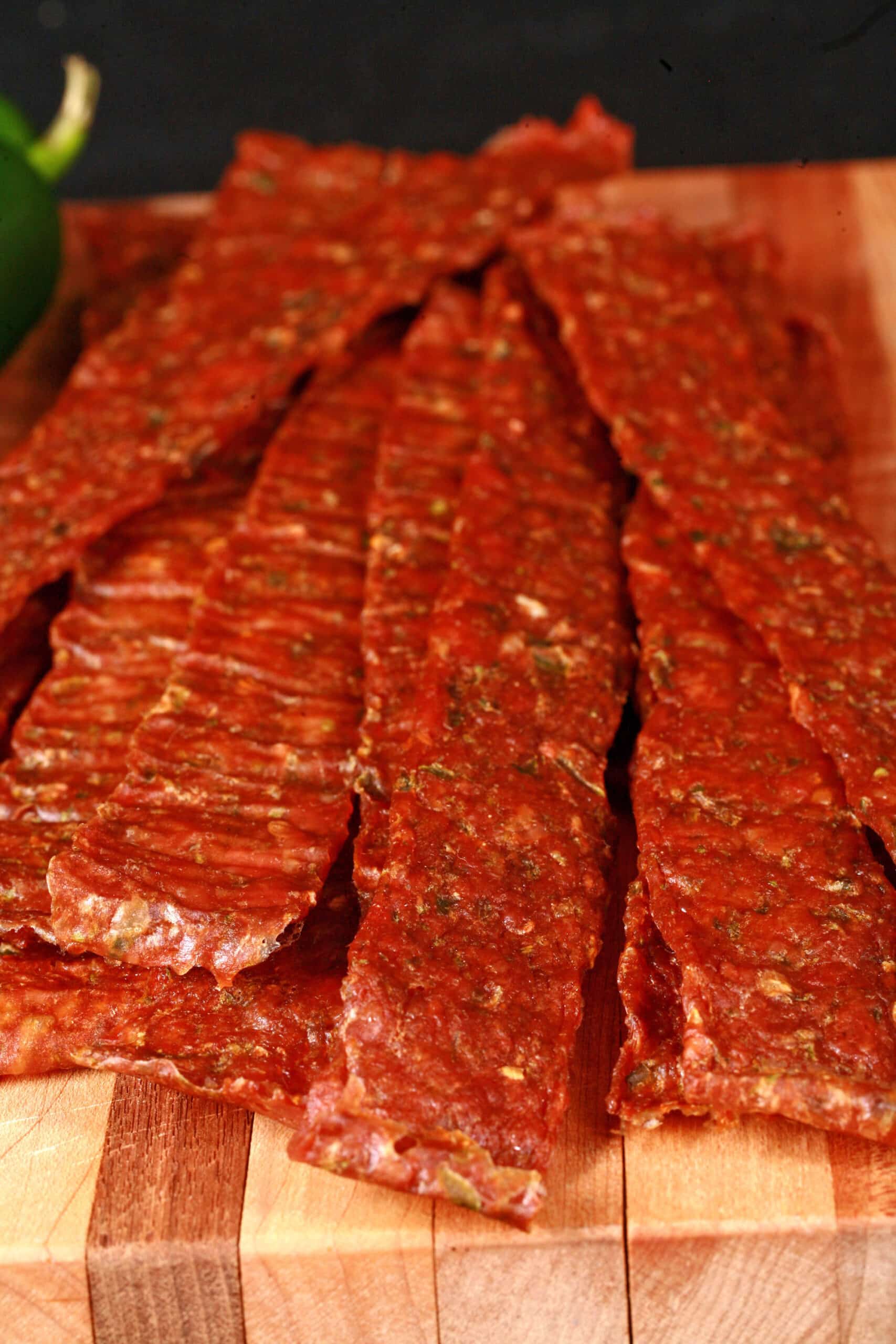 A pile of homemade chicken jerky on a wooden cutting board, along with 2 jalapenos.