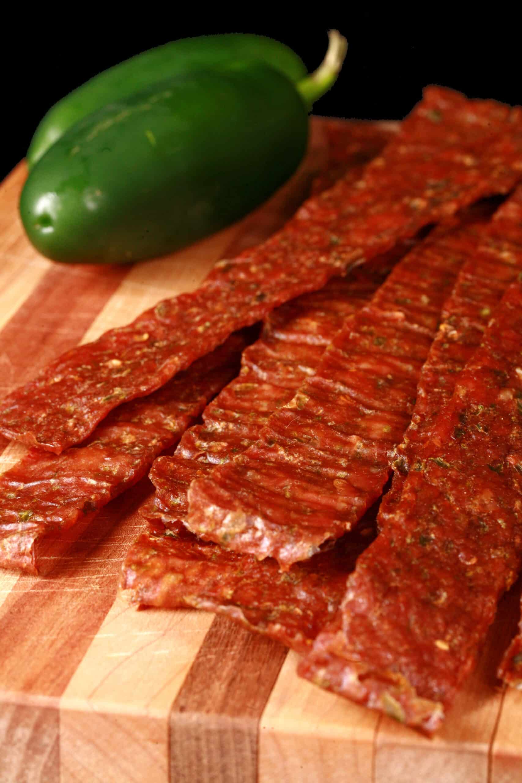 A pile of homemade jalapeno chicken jerky on a wooden cutting board, along with 2 jalapenos.