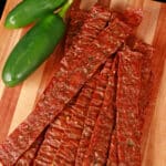 A pile of homemade jalapeno chicken jerky on a wooden cutting board, along with 2 jalapenos.