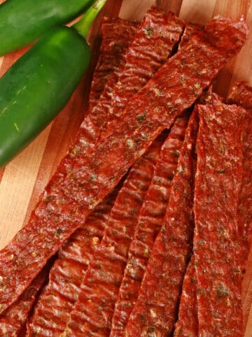 A pile of homemade jalapeno chicken jerky on a wooden cutting board, along with 2 jalapenos.