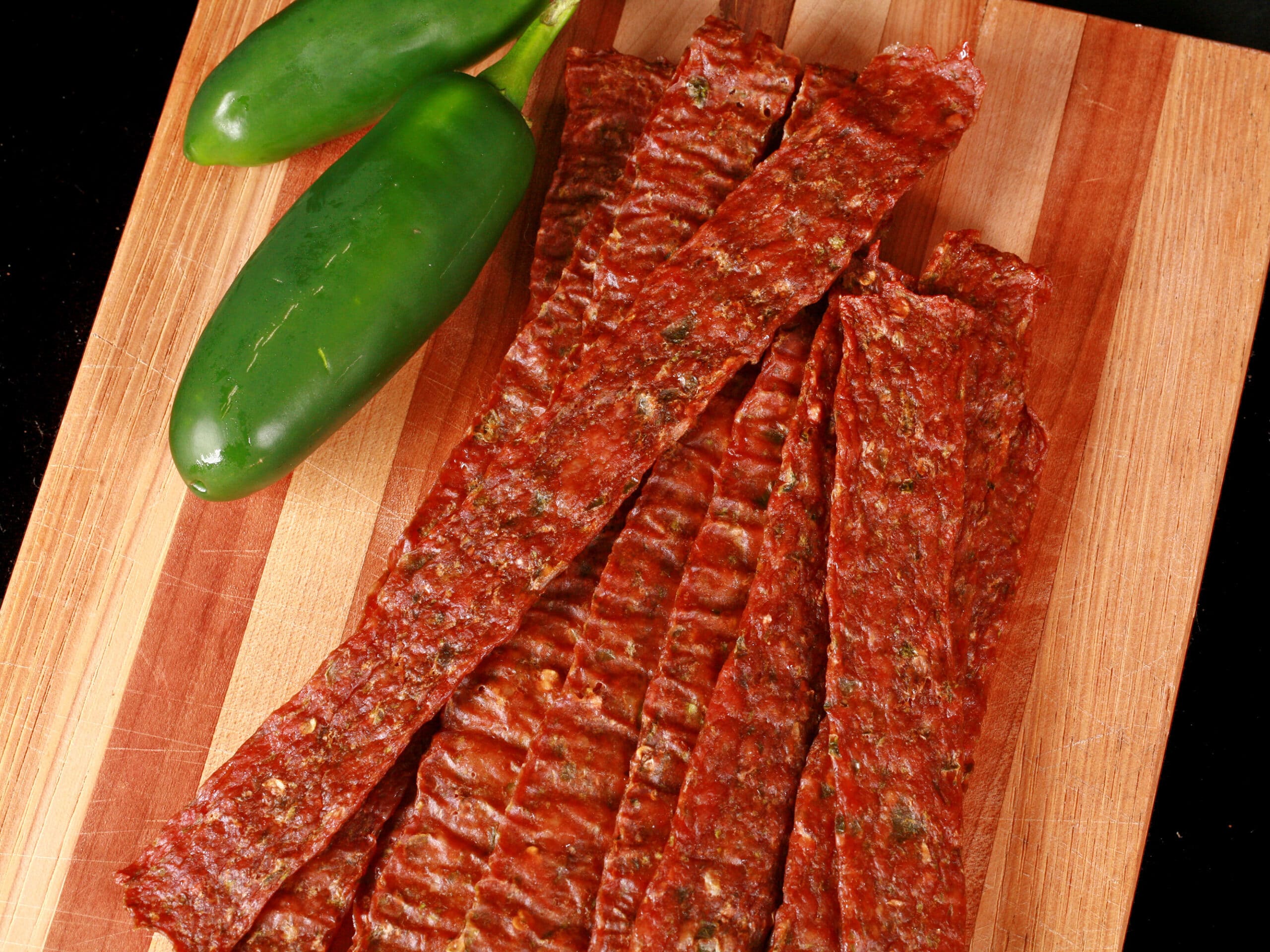 A pile of homemade jalapeno chicken jerky on a wooden cutting board, along with 2 jalapenos.