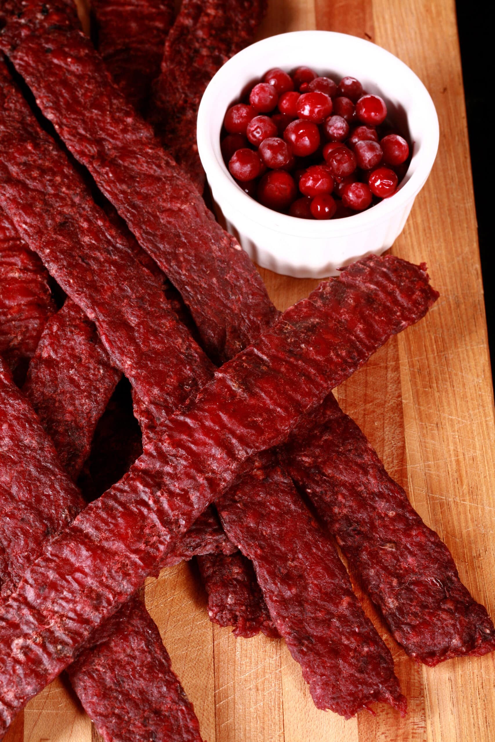 Several strips of homemade lingonberry elk jerky on a wooden cutting board, along with a small bowl of partridgeberries.