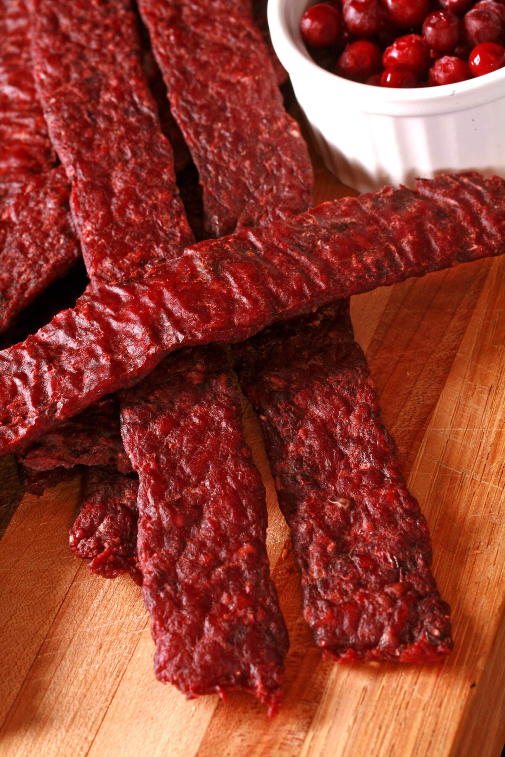 Several strips of homemade lingonberry elk jerky on a wooden cutting board, along with a small bowl of partridgeberries.