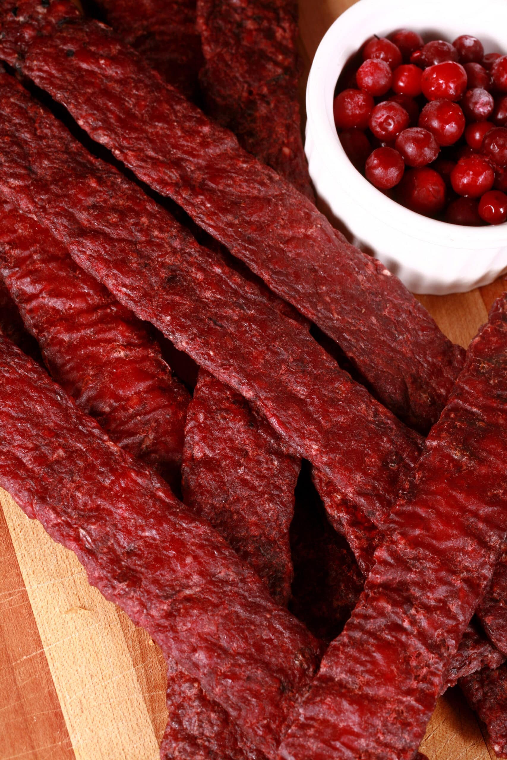 Several strips of homemade elk jerky on a wooden cutting board, along with a small bowl of lingonberries.