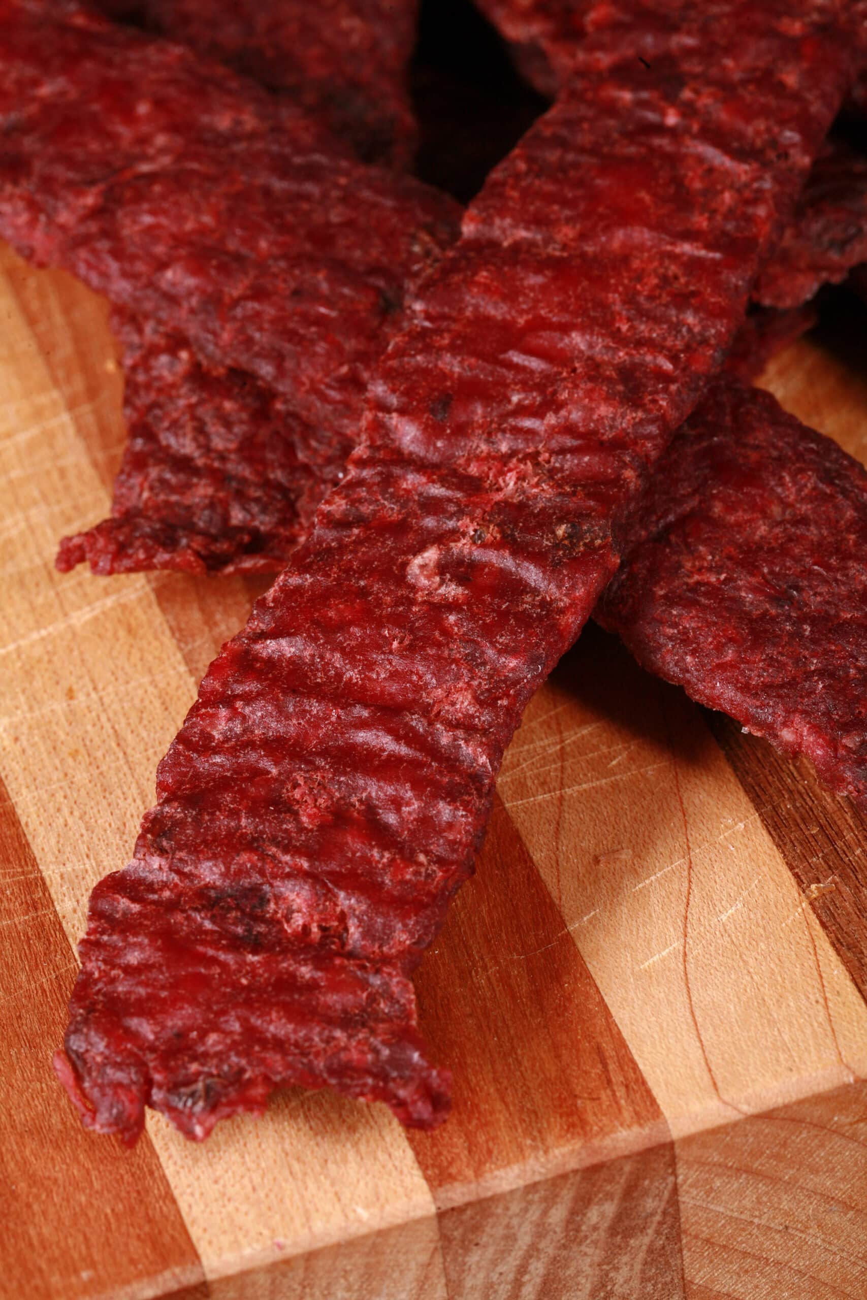 Several strips of homemade lingonberry elk jerky on a wooden cutting board, along with a small bowl of partridgeberries.