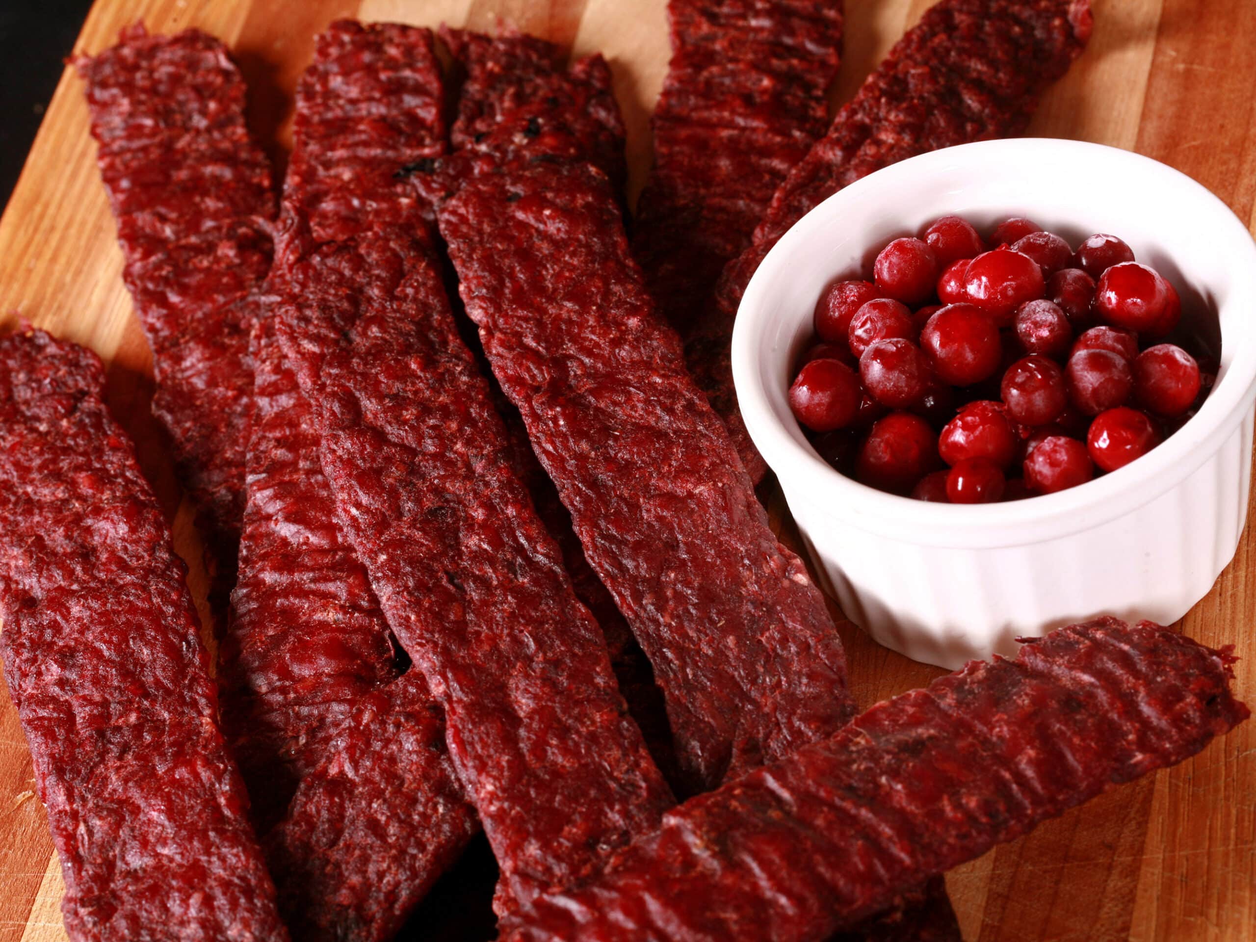 Several strips of homemade elk jerky on a wooden cutting board, along with a small bowl of lingonberries.