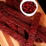 Several strips of homemade lingonberry elk jerky on a wooden cutting board, along with a small bowl of partridgeberries.