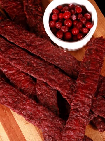 Several strips of homemade lingonberry elk jerky on a wooden cutting board, along with a small bowl of partridgeberries.